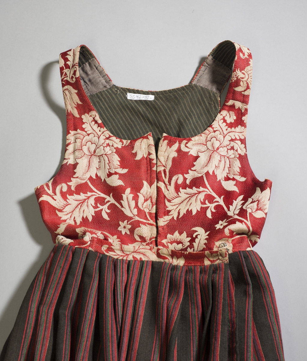 Håndsydd livkjole med stripete ullskjørt og rødt ullliv med store plantemotiv. Livet har rett hals bak, skjøtte seler og mangler ryggsøm. Lukningen består av fire par hekter og tre par hemper til snøring. Åpen klaff.
Fõr av stripete håndvevd ullstoff.
Nederst på skjørtet et børstebånd og skoning av et grindvevd bånd.
Skjørtet har en hengefald.