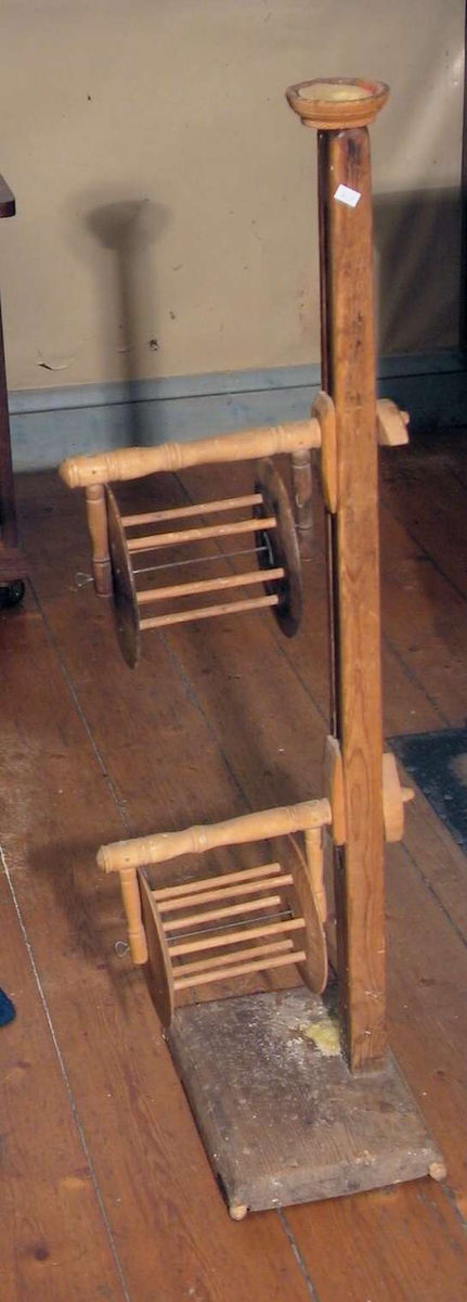 Garnvinde av tre. Den har to hjul festet til en høy stang. Stangen står på en rektangulær, flat fot.