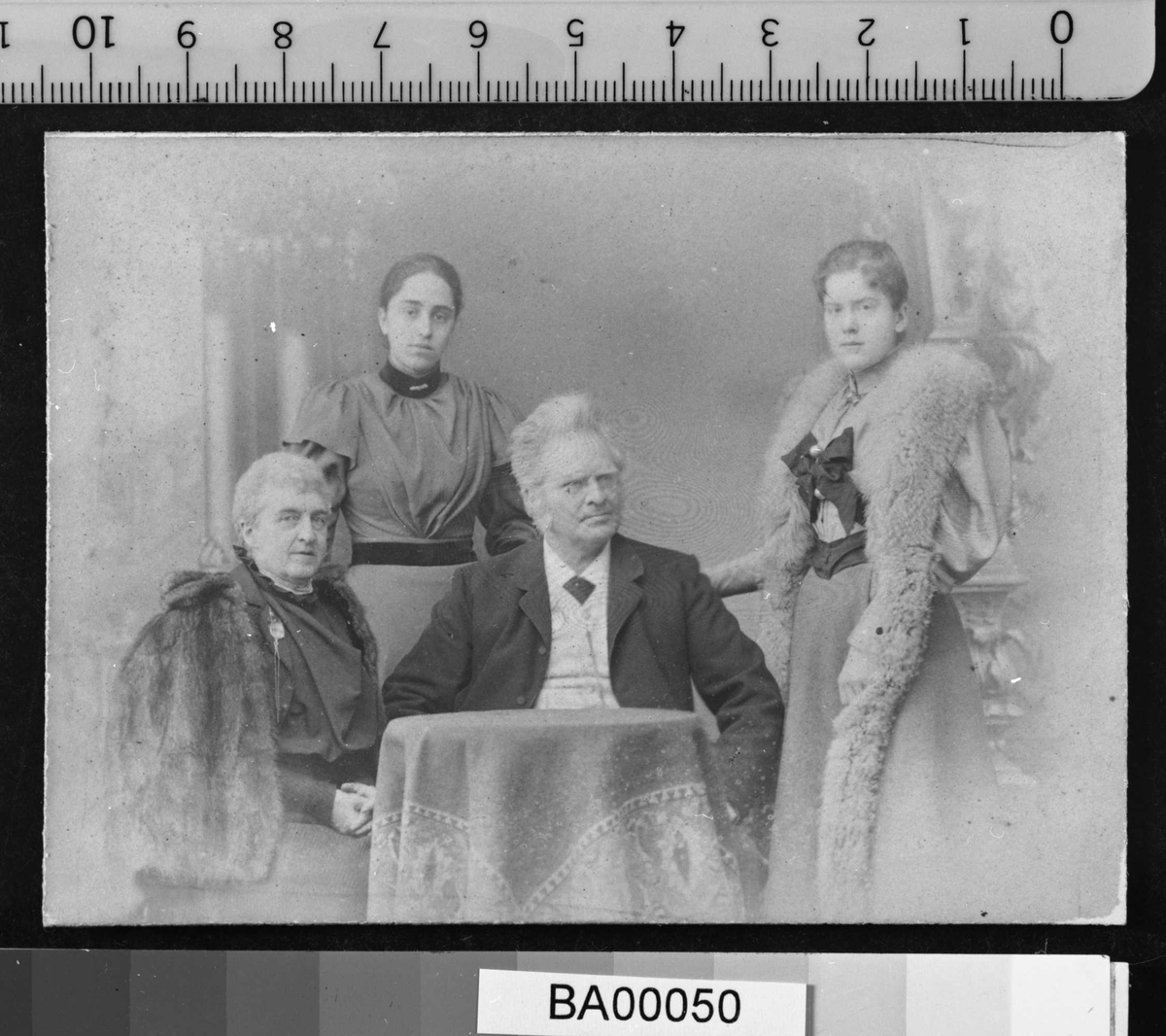 Atelierfotografi; bakerst står to jevngamle unge damer, den ene med lang pelsstola hengende ned langs med skjørtet og en stor mørk sløyfe bundet midt for brystet; den andre unge dame mer enkelt kledd i lys lang kjole med mørkt bånd rundt taljen og halsen isatt enkel brosje. Inn til et lite rundt bord sitter en dame med pelskape til høyre, og i midten en mann med henderne i siden, nesebriller, mørk jakke og slips, hvit skjorte og vest. Teppebakgrunnen viser antikke bygningsfragmenter. De tre damene ser direkte på betrakteren, mens mannen ser skrått til venstre.
