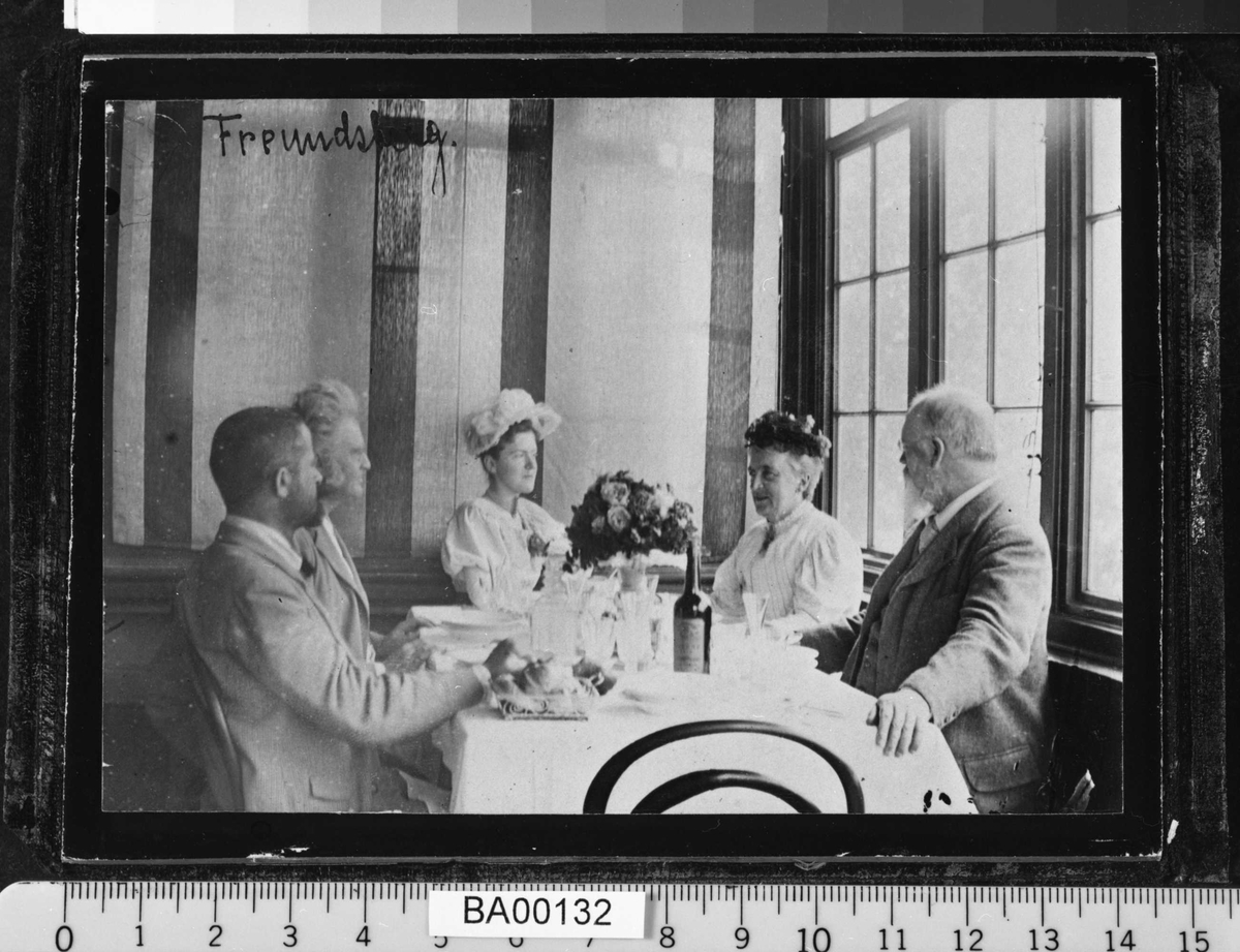 Fotografi fra spisesal; ved bordet sitter to damer og tre herrer; de ser på hverandre.