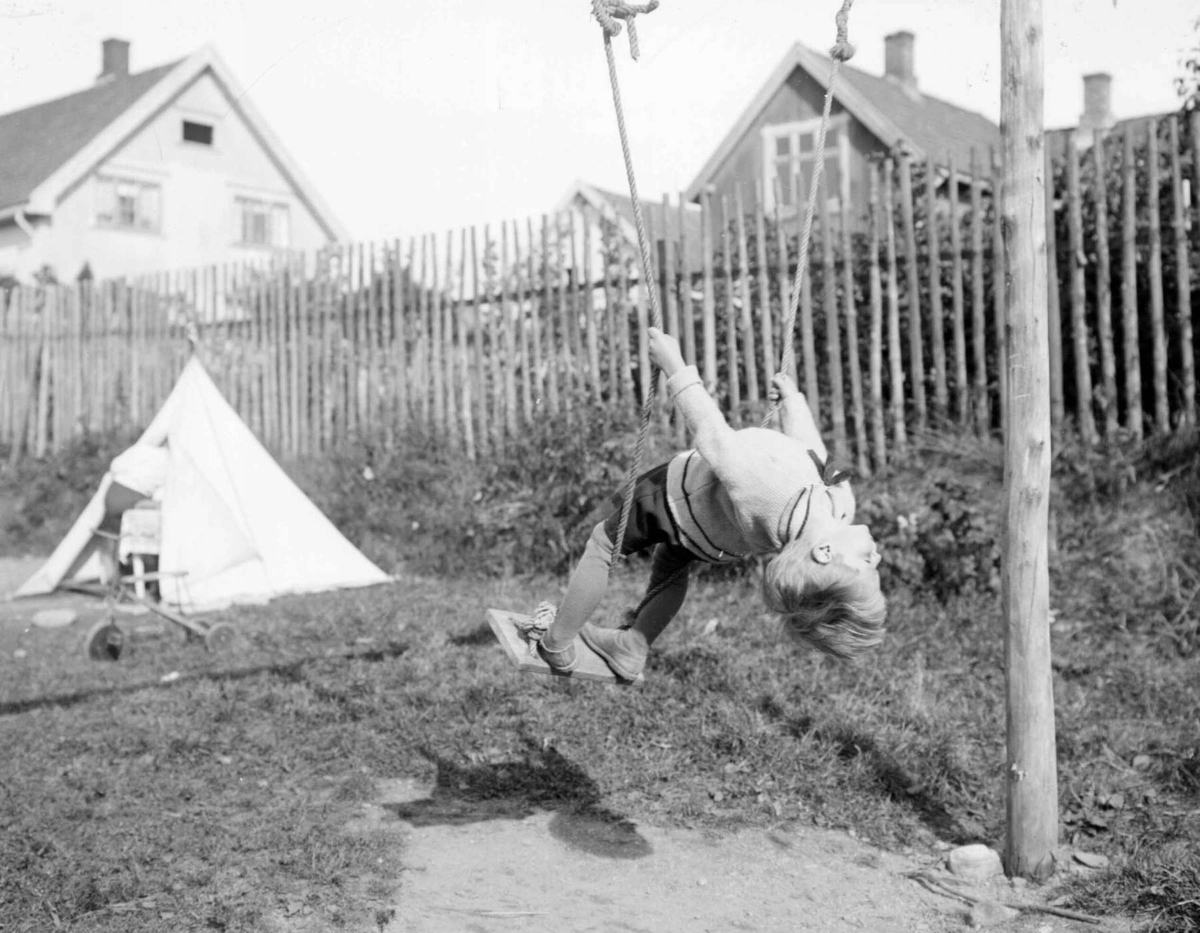 Liten gutt i huske. Gunnar Berge. Hus i bakgrunnen. Lite telt og tråsykkel.