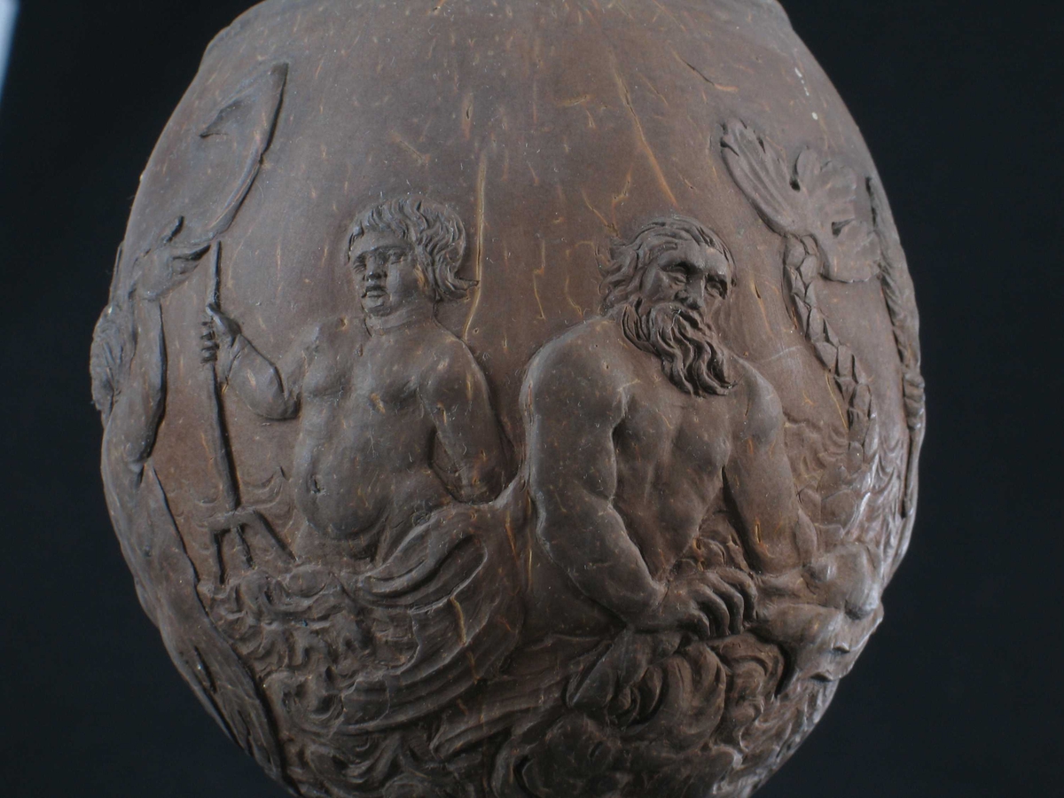 Foten har fire ornamentfelt i relieff, to av dem med blomsterdekor, de to andre med blad og overflødighetshorn. Pokalen bæres av en Neptunfigur utskåret i elfenben, med løftet venstrearm (denne har hatt scepter) og delfin mellom benene. På Neptuns hode en pokal av en kokosnøtt med toppen avskåret som lokk, og med beninnfatning.  Pokalen har relieffutsmykning med nereider og tritoner, som blåser i en koncha (basunlignende instrument). Lokket har fire relieffmotiver med nøtter og to med blomster.  Lokkgrep av ben, balusterformet, med sittende apekatt i metall på toppen.