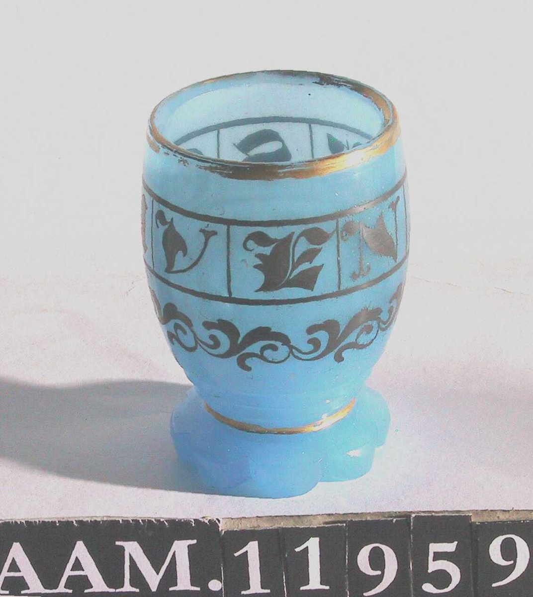 Beger  merket   SOUVENIR  Blått glass,   forgylt dekor.  Rosettformet bunn, lite tønneformet beger  med forgylt rand, tekst rundt med gotiske bokstaver: SOUVENIR.antag. Baccarat, eller annet fransk glassverk.  Tilstand: forgyllingen litt avslitt, ellers bra.
