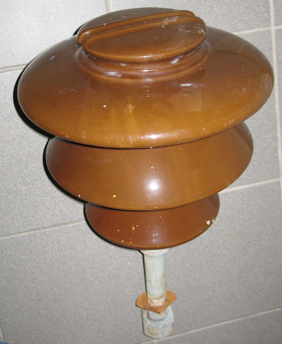Isolator til bruk i kraftlinjer. Festebolt i utspart senterhull i isolatoren. Bolten er gjenget i enden, og det medfølger en skive og en mutter. Brun farge, det finnes også grønne av samme type. Typen isolator vises på forsidebilde i boken Kraft og børs, Arendals fossekompani 1896 - 1996.  På den ene er rester av kabel .