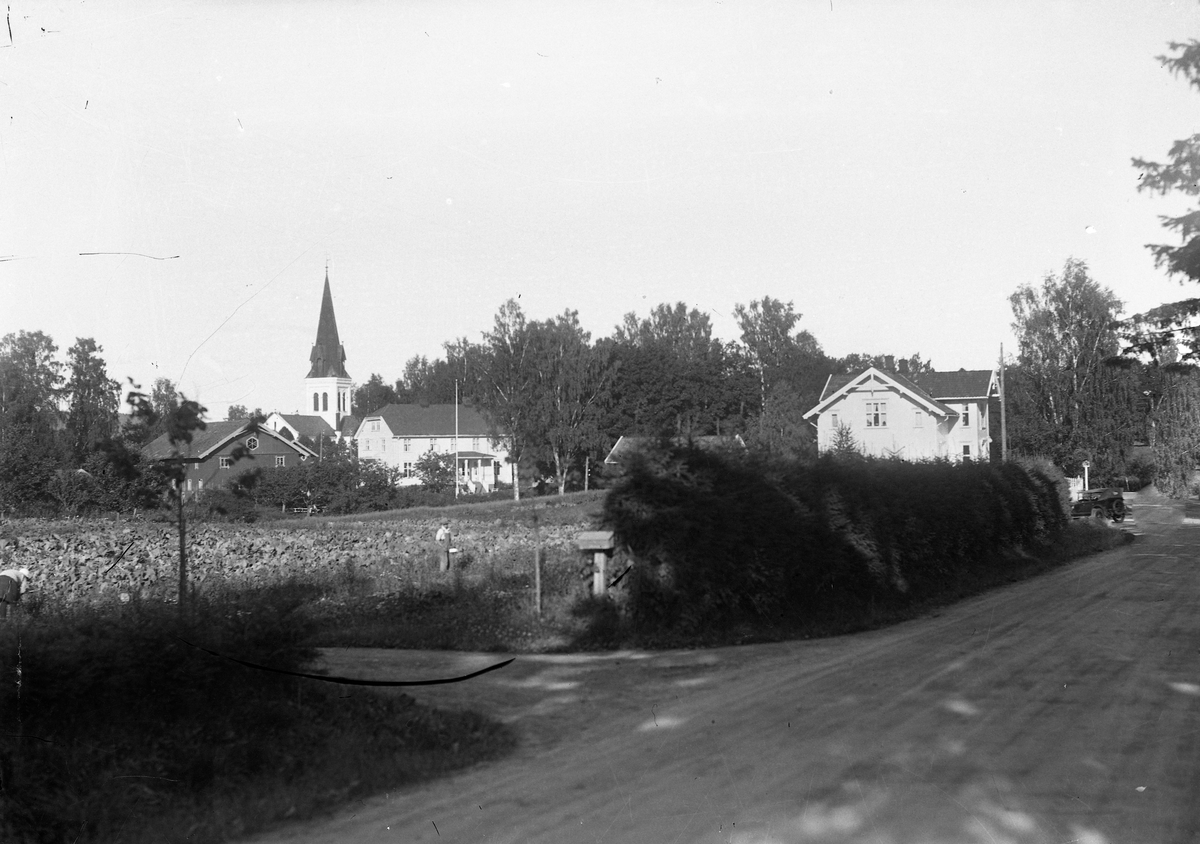 Området ved Eidsvoll Kirke. Huset til Haug/Flyen til høyre - med lensmannskontor. Mellomkrigs bilmodell på veien. Bildet må være etter 1922, da Eggerskjæringa var ferdig. Låven til lensmann Aasgård til venstre.