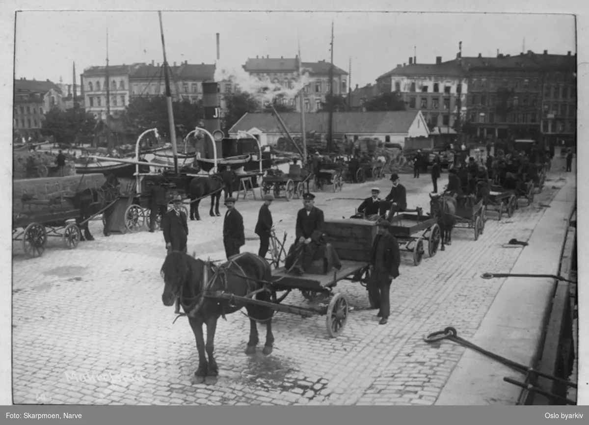 Havneliv, dampbåter, seilbåter, mange hest med kjerre, mange menn, lagerbygning på Tordenskjolds plass, masse trekasser utenfor, bygårdene fra tidligere Sjøgaten i bakgrunnen.