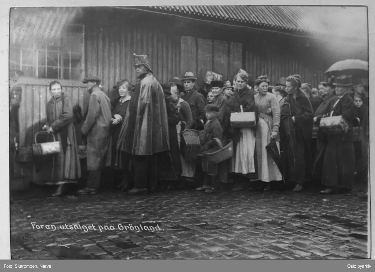Kø av mennesker utenfor utsalget på Grønland. Kvinner og menn, kvinner med kurv, mann med regnkappe.