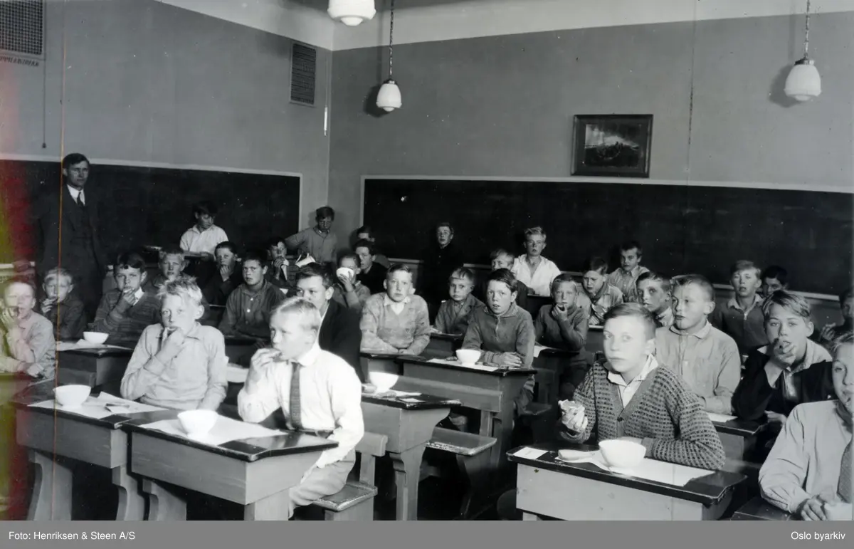 Skoleklasse. Skolefrokost i klasserommet. Elevene ved pultene med mat og melkeflasker. Oslofrokost.