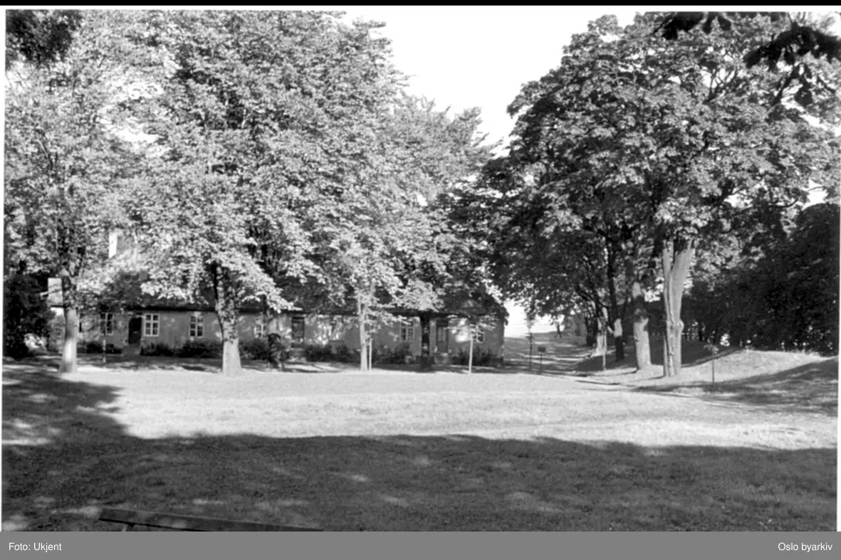 Festningsområdet (Lensherrens hage) på Prins Carls bastion og nordover mot hovedvakten og borgporten mot øst og Kongens gate. Barakkene (1747) ses midt i bildet. Munkedammen ble gjennskapt i 1960. Gardist marserer fra porten til hovedvakthuset i bakgrunnen.