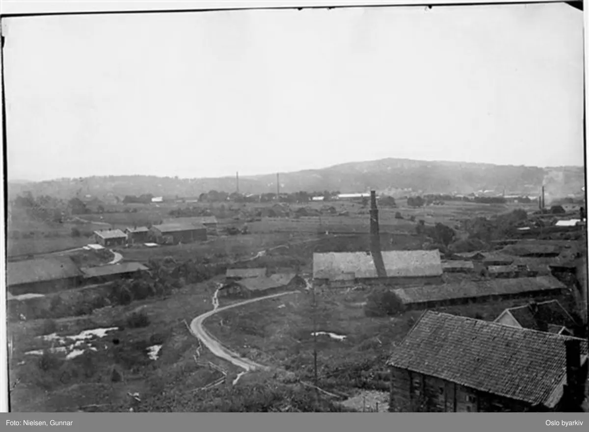 Fotografert mot sørøst fra taket på Østre Hasle gård i Økernveien. Økernveien i forgrunnen til høyre. Låven som står omtrent hvor inngangen til Hasle T-banestasjon nå ligger, sto til langt ut på 1950-tallet. Veien som går i sving var den daværende forbindelse mellom Økernveien og Hovinveien. Til venstre Hovin teglverk. De små husene til høyre kan være Hasle teglverk. De to pipene midt på i bakgrunnen tilhører fyrstikkfabrikken på Grønvold. Den rykende pipa til høyre tilhører Ensjø teglverk, omtrent der Ensjø T-banestasjon ligger i dag. Mer detaljert informasjon om motivet finnes.