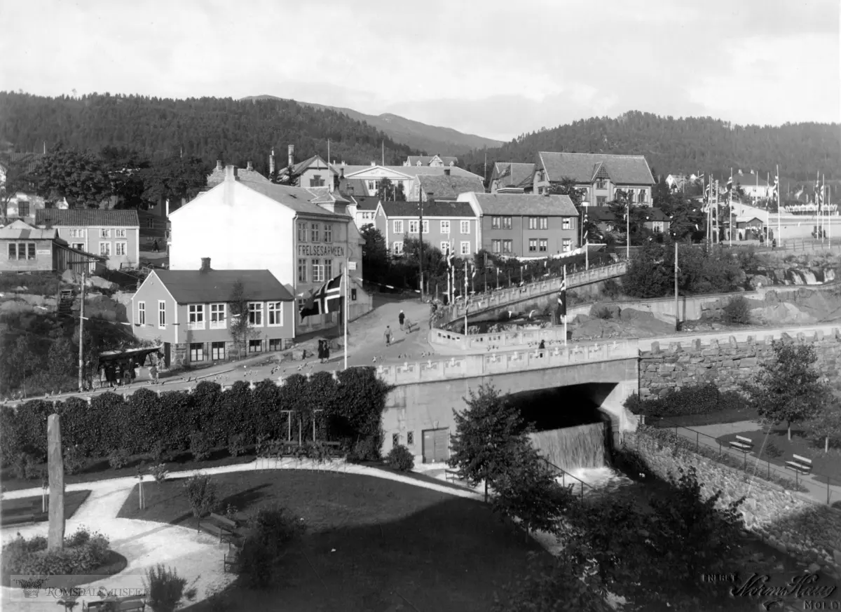 Elvebakken, Romsdalsgata med elva og broen sett fra Gotfred Lies. Frelsesarmeen. 1931. "Vegen opp Elvegata (no Spolertbakken) til Skuleplassen var pynta med flagg. l framgrunnen Gotfred Lies plass. (Foto Norm. Haug)"