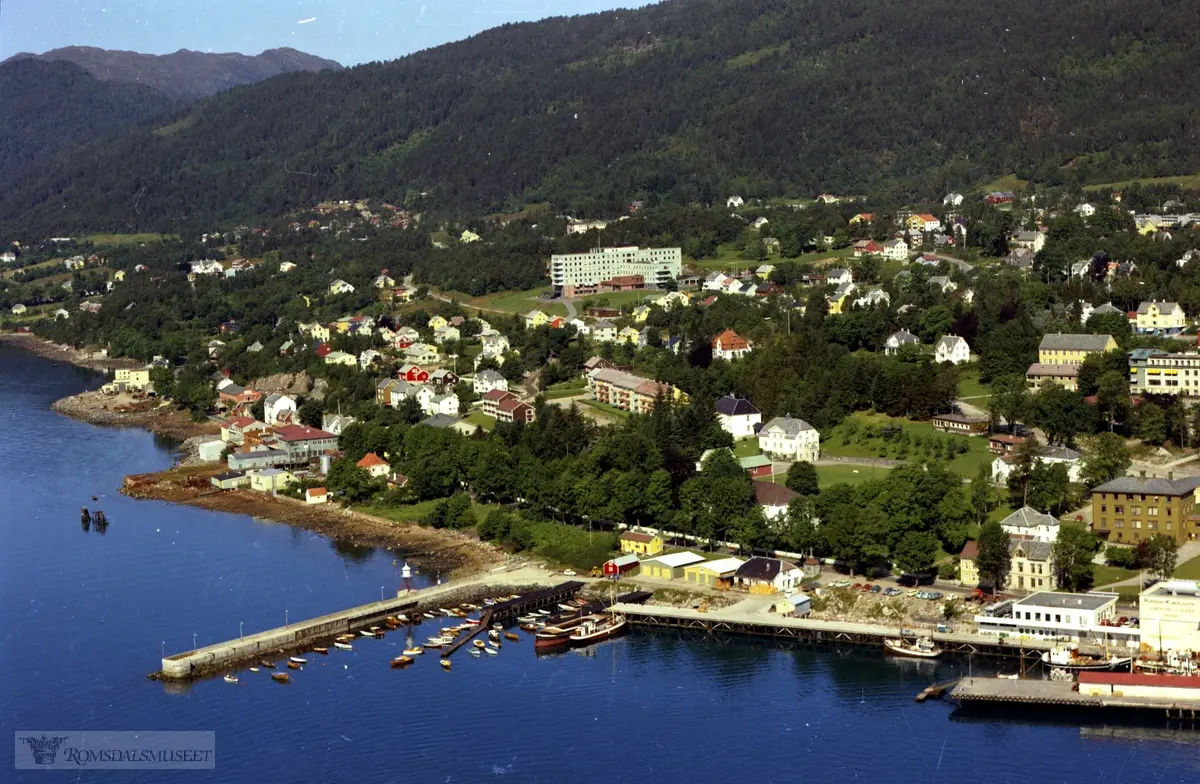 Julsundvegen med Reknesmoloen..Fjærestua (Lassenfamiliens hus) ligger i skogen til venstre, ved sjøen. Flere hvite hus midt i bildet tilhørte Reknes Sanatorium. .I vest ser vi Christie & Opsahl verkstedet, gult bygg. Det røde bygget på øversiden av Julsundveien er "Gamle" Heimdal. Bygget ble overtatt av Wiigs Maskin. Den grønne bygningen på nedersiden av Julsundveien er Heimdalmotor A/S's bygning. Den bygningen ble revet i forbindelse med bygging av stadion. Parkeringsplassen vest for Stadion er hvor bygget stod.