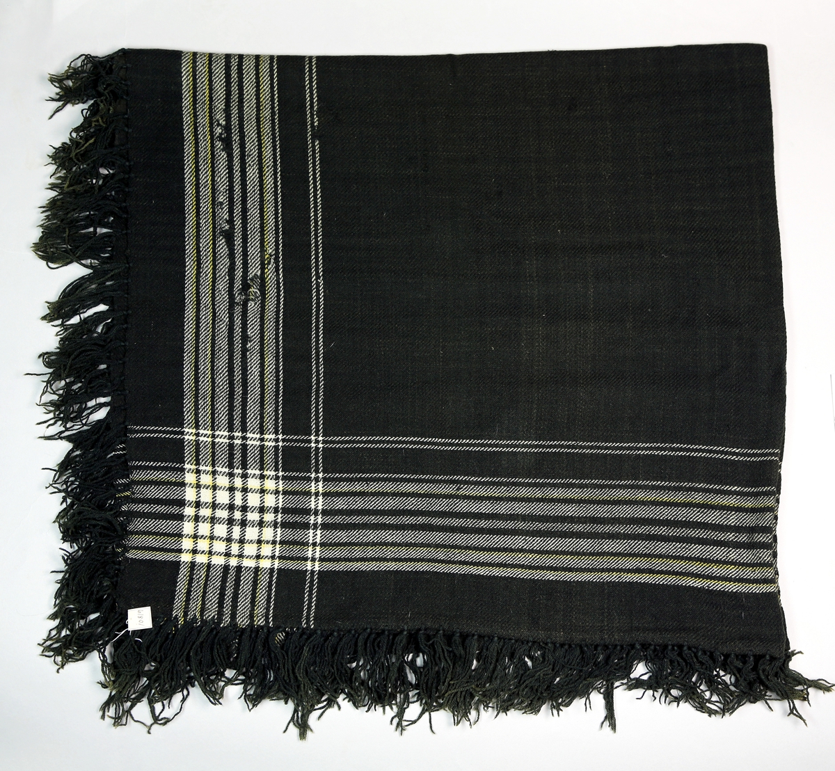 Sjal i ull med frynser på alle kanter. Det er vevd inn mønster bestående av lyse render langs kantene. Det er skjøtt sammen av to stykker, og skjøten er håndsydd og er omtrent på midten. 