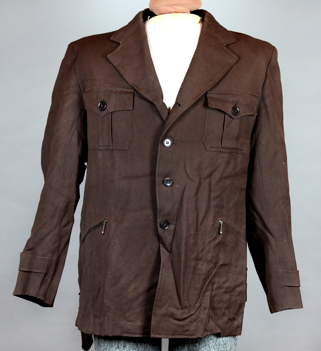 Dress bestående av jakke og bukse. Jakka er enkelt spennet og kneppes med tre knapper, og i tilleggg er det et belte rundt livet. Buksa er kort, - knekort. Jakka har fire lommer utenpå, hvorav de to nedre kan lukkes med glidlås og de to øvre lukkes med knapper. Den er fôret med silke. Buksa har både plastknapper og metallhekte. Nede kan buksa strammes med stropper rundt leggene. 