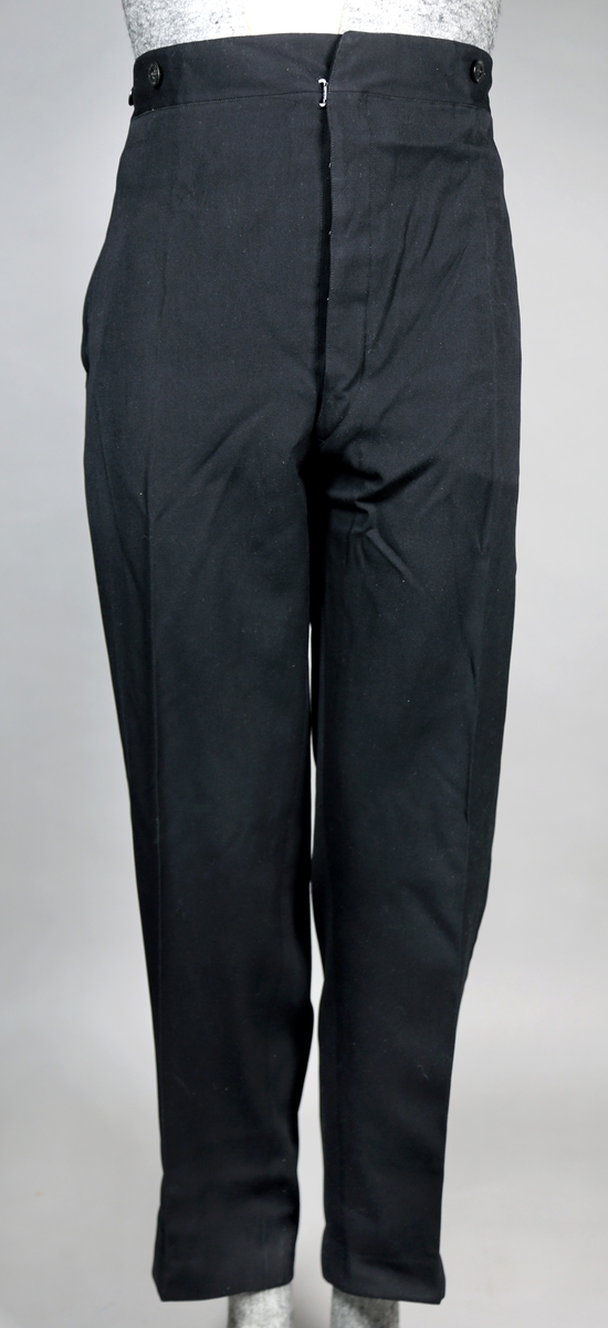 Svart dress med vest og lang jakke. Jakka er dobbeltspennet og kneppes med fire knapper, som er trukket. Jakka har splitt bak. Den har silkefôr, bortsett fra i ermene. Buksa har både plastknapper og metallhekte, og tre lommer. 