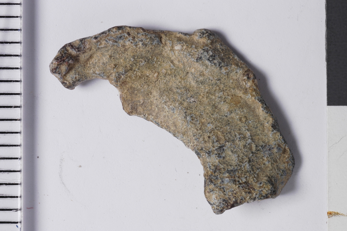 Del av plakett fra vareplombe, som har hatt to nagler. Gjenstanden har tegn til preg i form av perlerand i ytterkant. 