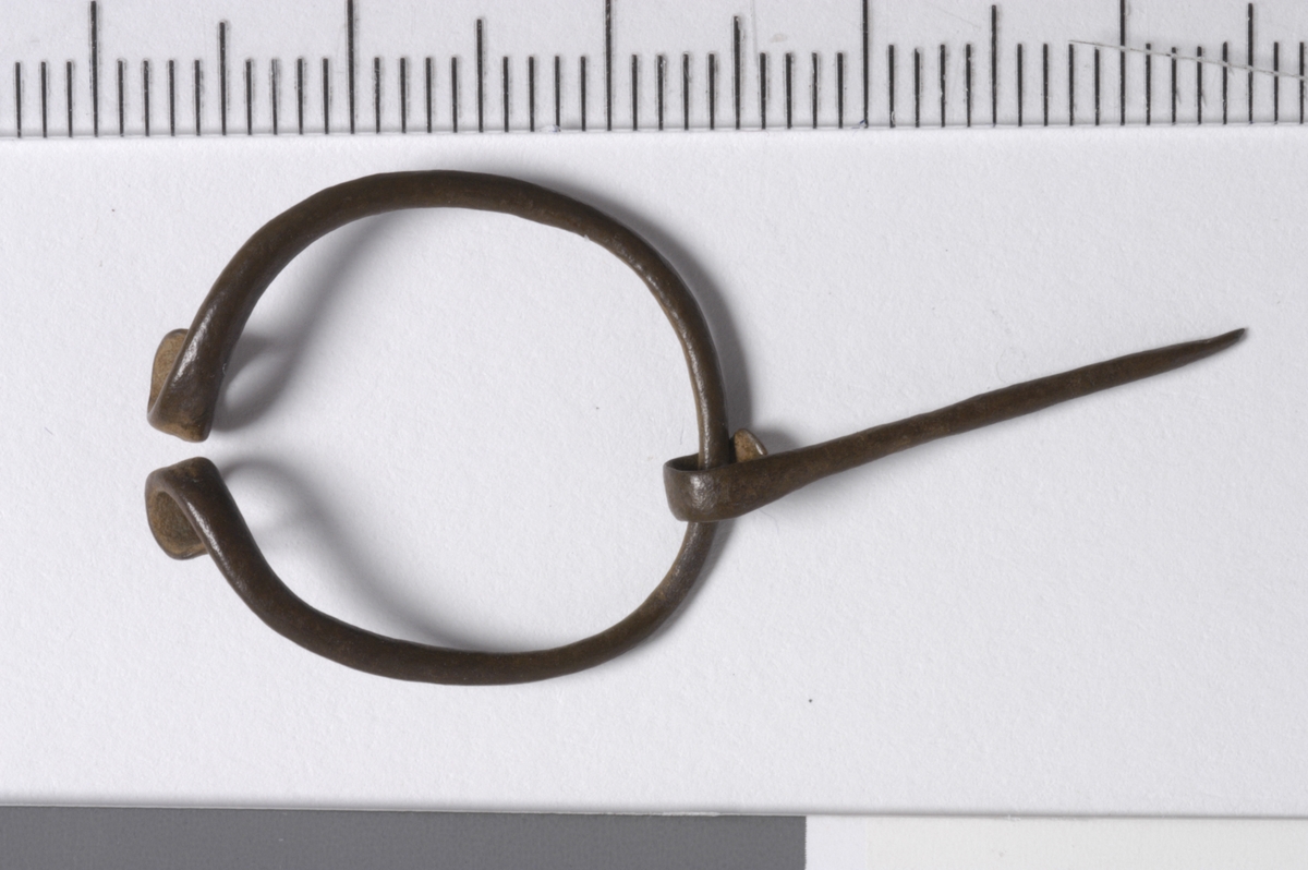 MALV: DIAM. 20  T. 1 MM, ÅPEN RING HVOR ENDENE ER OPPRULLET. UTEN DEKOR. TORNEN ER 23, 2 MM LANG MED STØRSTE BREDDE 2 MM. IDENTISK MED "HESTESKO-SPENNEN" FIG. 14 S. 131 (BLOMQUIST R. ) SOM VAR VANLIG PÅ 1000-TALLETOG FREMOVER. BRUKT TIL Å FESTE SAMMEN SKJORTEN I HALSÅPNINGEN. ETTER1000-TALLET ER RINGEN VANLIGVIS SLUTTET. 