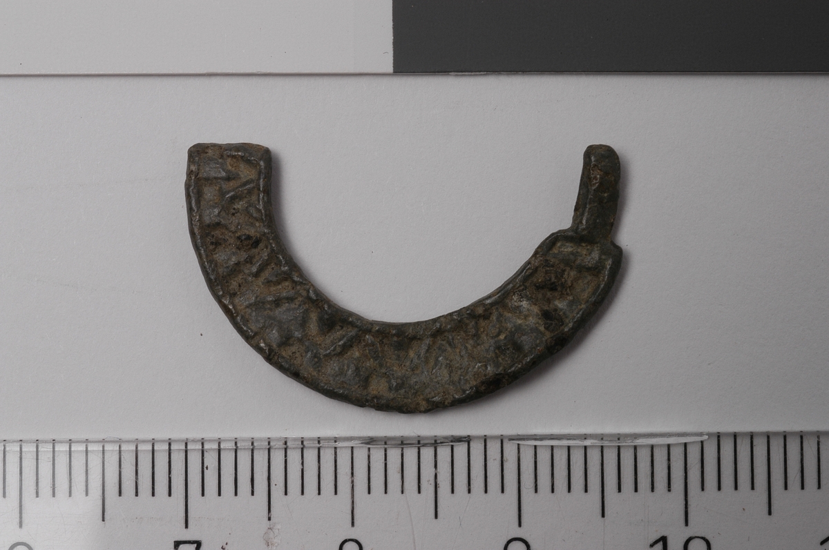MALV: DIAM. 27  RAMMENS B. 5 MM, BÅNDFORMET. DEN ENE SIDEN HAR DEKOR, MEN VANSKELIGÅ TYDE, STØPT. SER UT SOM DEFORMERTE BOKSTAVER. LIGNER SPENNEN FRA LUND- BLOMQUIST L. 1947: BILD 22, 5. DATO: 1300-TALLET. 