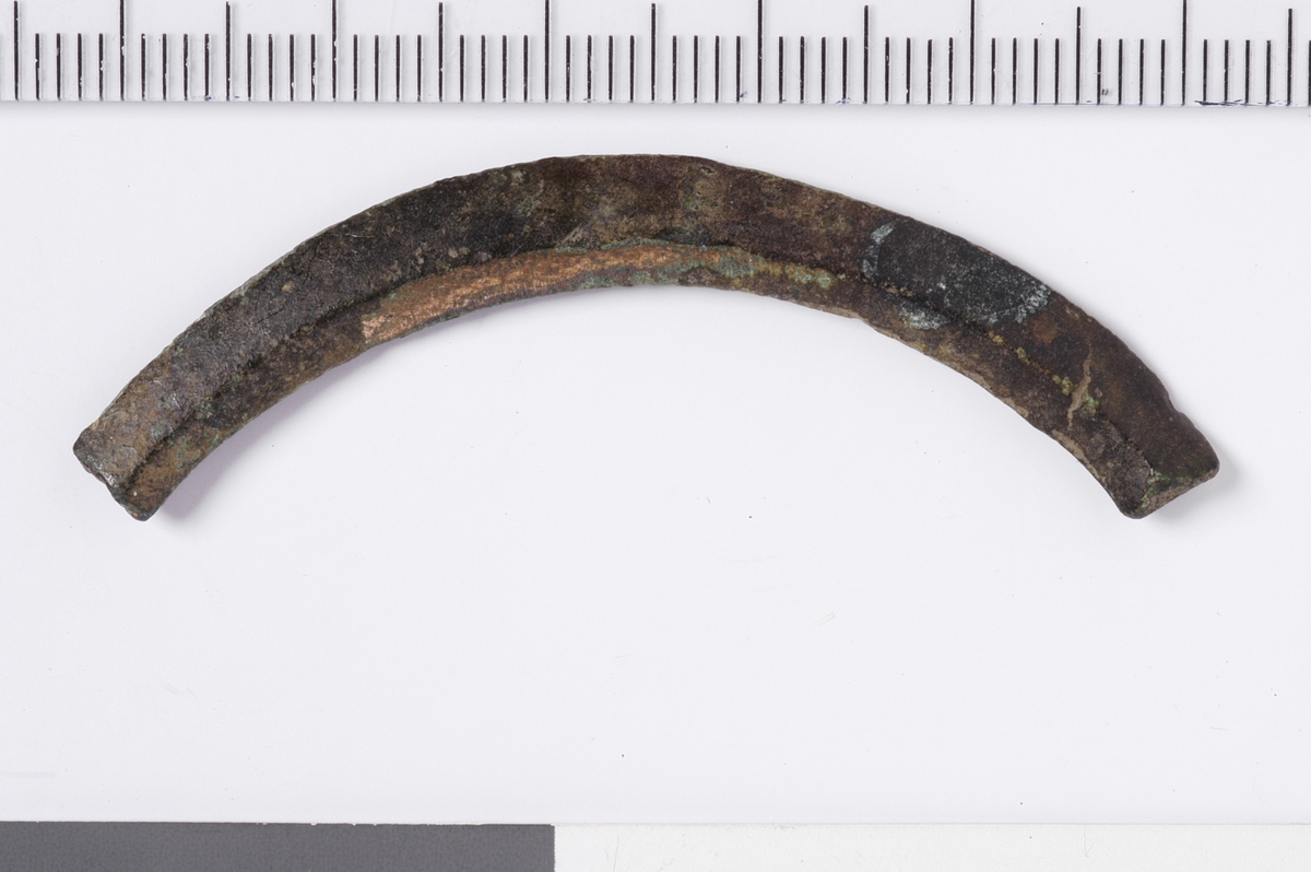 Et lengre og buet metallstykke med bruddflater i begge ender og trapesformet tverrsnitt. Gjenstanden er uten dekor, men støpt og filt. På undersiden er det forsatt noe støpespor som antyder at gjenstanden ikke er ferdig men et emne. Dette kan være en del av et emne til en ringspenne.