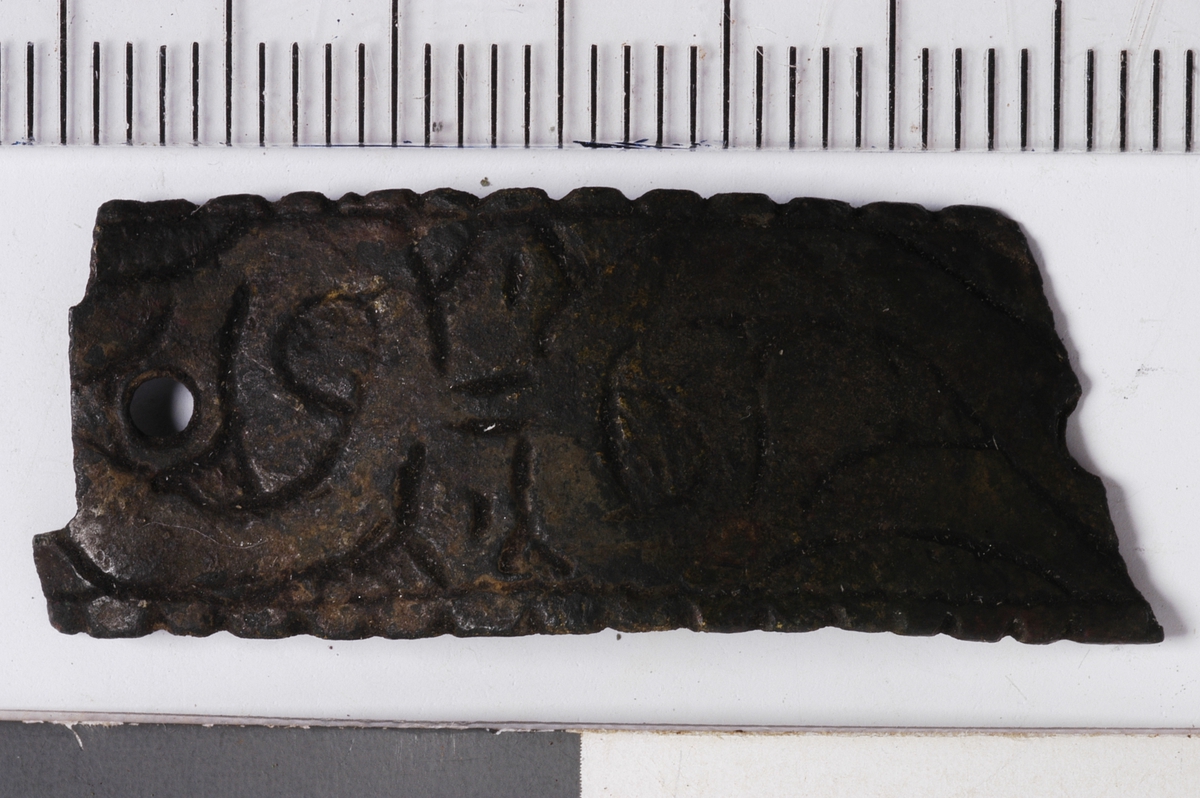 MALV: L. 33 MM B. 12 MM, BÅNDFORMET PLATE, AVBRUKKET I BEGGE ENDER. ET HELTNAGLEHULL OG ET AVBRUKKET. DEKORERT MED STILISRERTE PLANTEORNAMENTEROG BØLGEKANT. 