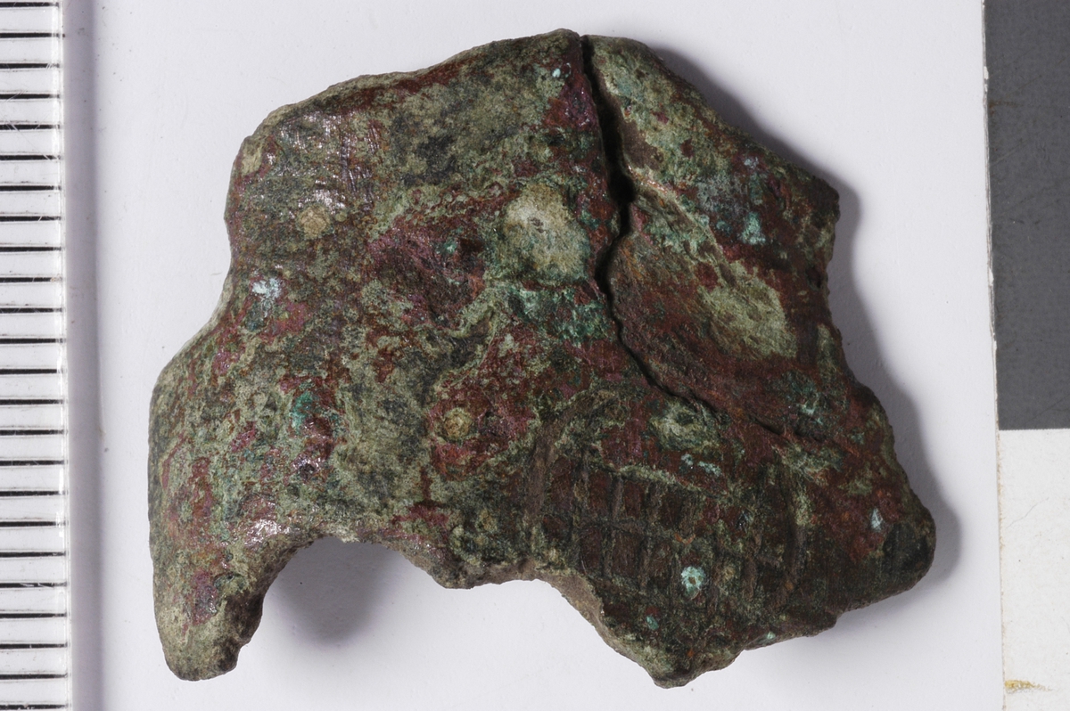 MALV: ST. B. 25 MM TYKKELSE 2, 5 MM, BØYD, UJEVN PLATE, UTEN NOEN DEFINERT FORM. PÅ PLATENSYNES DELER AV ET ORNAMENT I FORM AV EN LITEN SIRKEL MED ET OPPRUTETFELT TVERS OVER. 
