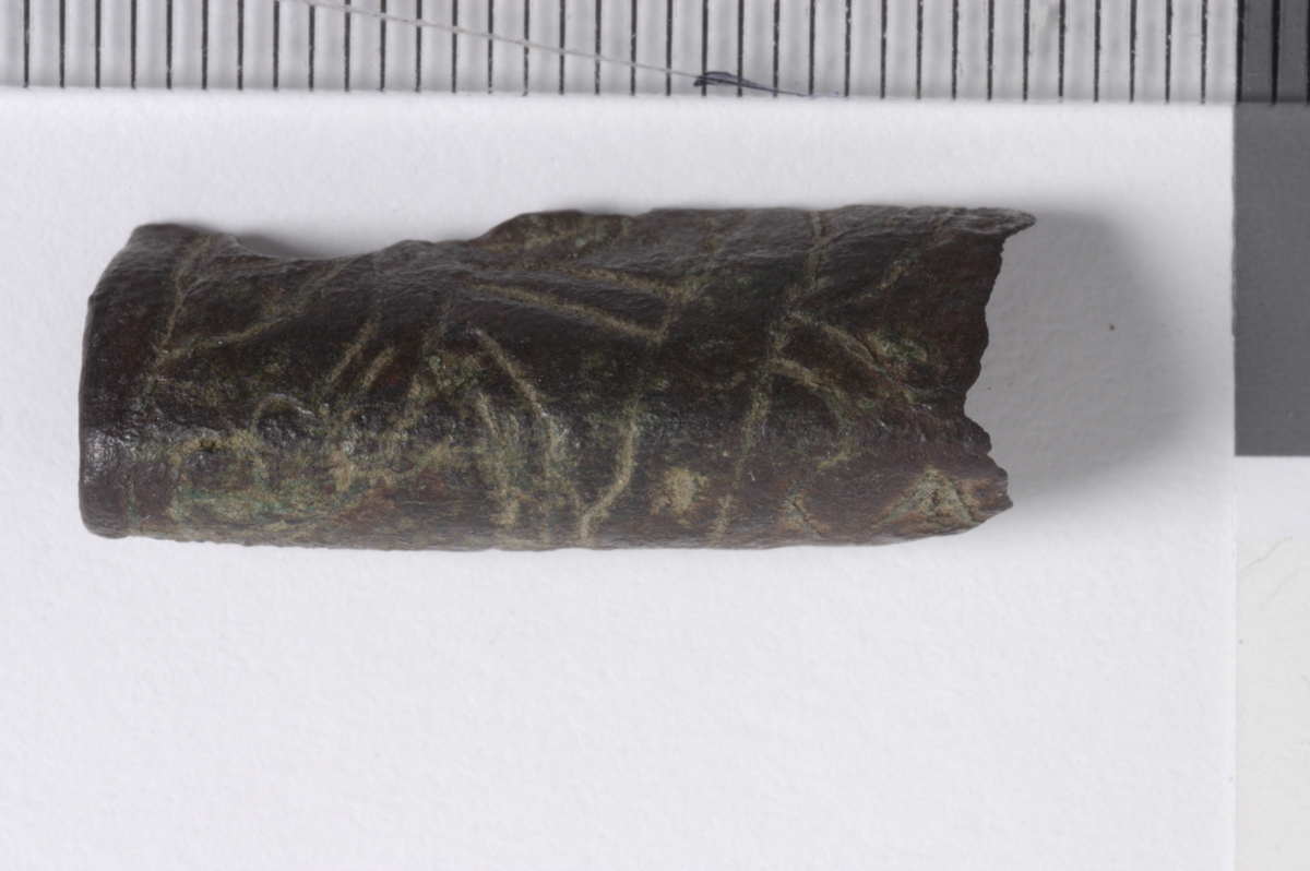 MALV: L. 25 MM, BÅNDFORMET FRAGMENT SOM ER BØYET. OVERSIDEN HARINNPRESSET DEKOR. DEN ENE ENDEN BEARBEIDET. 