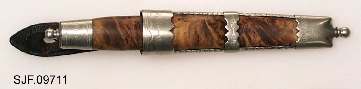 Tollekniv med slire.  Slira er signer "T. B. ", som skal være initialene til Trygve Brenna fra Åmot på Modum.  Bladet er 8, 2 centimeter langt og 1, 9 centimeter bredt ved brystinga.  På den bakre delen av nakken, som er om lag 0, 5 centimeter bred, er det filt to tverrstreker og et kryss.  Høyre bladside har tre innstemplete initialer, antakelig "Ø. L. S. ", innskrevet i ei buet ramme.  Denne signaturen ble brukt av eggsmeden Knut Knutsen Øyo (1860-1921), og seinere av firmaet til sønnene Lars, Thomas, Knut og Olav Øyo, «Øyo ljåsmie».  Dette navnet ble brukt etter 1. verdenskrig.  Firmanavnet ble for øvrig endret til «Brødrene Øyo» i 1941, som en konsekvens av at produktspekteret bedriften kunne levere var blitt bredere, og av at ljåblader ikke lenger var det dominerende produktet.  Bladet på denne kniven bærer preg av å være slipt mange ganger. 

Skaftet er lagd av flammebjørk Det er snaut 9, 5 centimeter langt og har en noe konveks form i lengderetningen og et ovalt tverrsnitt (snaut 2, 5 X 1, 95 centimeter på midten).  Begge ytterender er forsterket med 2, 0 centimeter brede nysølvholker med inngraverte linje-, bue- og rombemønstre.  I enden av bakholken er det påsatt ei rombeformet plate, som den dreide knoppen enden av knivbladets tange er forankret i er festet mot.  Tredelen av skaftet har en lys gulbrun farge som muligens skyldes forsiktig beising.  Kniven - inkludert blad, skaft og knopp - er 18, 7 centimeter lang.  

Kniven har treslire, lagd av flammebjørk og bundet sammen av nysølvbeslag.  Slirebeslaget består av to spenger langs sidene, som holdes ihop av tre holker: en munningsholk (øverholken), en midtholk og en doppsko.  Munningsholken har samme dekor som holkene på skaftet, altså linjemønster (parallelt med overkant), bue- og rombemønster (skravert).  Nederkanten på denne holken har et profilert mønster på den sida som skulle vendes fram når kniven ble båret i bukselinninga.  På baksida er det påloddetei hengsle for en beltestropp med "smikk" av brunsvart lær.  Den bakre delen av denne stroppen har spalte for på- og avknepping, den fremre et hull for forankring av kniven i knoppen i skaftenden.  Midtholken på slira har inngravert to "skraverte" romber, og har profilerte kanter på forsida av slira.  Baksida er glatt, bortsett fra at det er her knivmakeren har gravert inn initialene sine med ei kort pil på hver side.  Doppskolen er tilnærmet rektangulær og har en trefliket "rombe-rosett" som en profilert overkant som dekor på forsida.  I enden av doppskoen er det påsatt en dreid knopp av samme type som den som er brukt i øverenden av skaftet. 

Redskapet veier 135 gram, herav sjølve kniven 70 gram. 