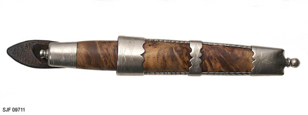 Tollekniv med slire.  Slira er signer "T. B. ", som skal være initialene til Trygve Brenna fra Åmot på Modum.  Bladet er 8, 2 centimeter langt og 1, 9 centimeter bredt ved brystinga.  På den bakre delen av nakken, som er om lag 0, 5 centimeter bred, er det filt to tverrstreker og et kryss.  Høyre bladside har tre innstemplete initialer, antakelig "Ø. L. S. ", innskrevet i ei buet ramme.  Denne signaturen ble brukt av eggsmeden Knut Knutsen Øyo (1860-1921), og seinere av firmaet til sønnene Lars, Thomas, Knut og Olav Øyo, «Øyo ljåsmie».  Dette navnet ble brukt etter 1. verdenskrig.  Firmanavnet ble for øvrig endret til «Brødrene Øyo» i 1941, som en konsekvens av at produktspekteret bedriften kunne levere var blitt bredere, og av at ljåblader ikke lenger var det dominerende produktet.  Bladet på denne kniven bærer preg av å være slipt mange ganger. 

Skaftet er lagd av flammebjørk Det er snaut 9, 5 centimeter langt og har en noe konveks form i lengderetningen og et ovalt tverrsnitt (snaut 2, 5 X 1, 95 centimeter på midten).  Begge ytterender er forsterket med 2, 0 centimeter brede nysølvholker med inngraverte linje-, bue- og rombemønstre.  I enden av bakholken er det påsatt ei rombeformet plate, som den dreide knoppen enden av knivbladets tange er forankret i er festet mot.  Tredelen av skaftet har en lys gulbrun farge som muligens skyldes forsiktig beising.  Kniven - inkludert blad, skaft og knopp - er 18, 7 centimeter lang.  

Kniven har treslire, lagd av flammebjørk og bundet sammen av nysølvbeslag.  Slirebeslaget består av to spenger langs sidene, som holdes ihop av tre holker: en munningsholk (øverholken), en midtholk og en doppsko.  Munningsholken har samme dekor som holkene på skaftet, altså linjemønster (parallelt med overkant), bue- og rombemønster (skravert).  Nederkanten på denne holken har et profilert mønster på den sida som skulle vendes fram når kniven ble båret i bukselinninga.  På baksida er det påloddetei hengsle for en beltestropp med "smikk" av brunsvart lær.  Den bakre delen av denne stroppen har spalte for på- og avknepping, den fremre et hull for forankring av kniven i knoppen i skaftenden.  Midtholken på slira har inngravert to "skraverte" romber, og har profilerte kanter på forsida av slira.  Baksida er glatt, bortsett fra at det er her knivmakeren har gravert inn initialene sine med ei kort pil på hver side.  Doppskolen er tilnærmet rektangulær og har en trefliket "rombe-rosett" som en profilert overkant som dekor på forsida.  I enden av doppskoen er det påsatt en dreid knopp av samme type som den som er brukt i øverenden av skaftet. 

Redskapet veier 135 gram, herav sjølve kniven 70 gram. 
