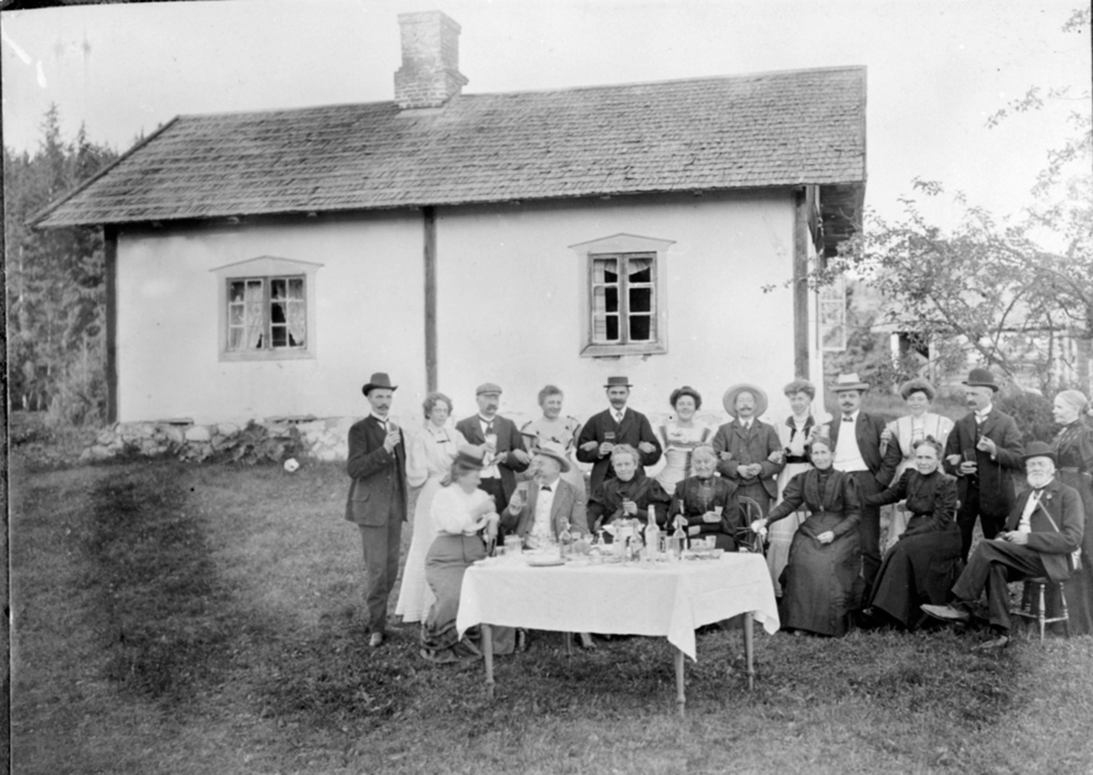 Festlig lag med mye folk utendørs, bord med flasker og glass. Rappet tømmerbygning.