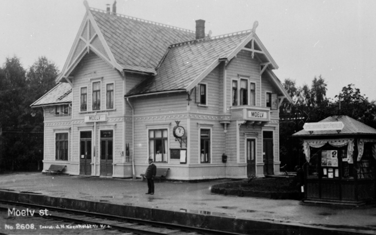 Postkort, Ringsaker, Moelv stasjon, jernbanestasjon med Narvesen kiosk, stasjonsmester på perrongen, stasjonsbygningen har fått panel og malt i en lys farge,