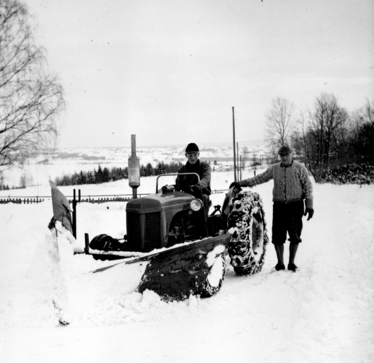 Traktor med snøplog i Lodviken, Helgøya. Bård Dobloug kjører traktoren, Mattis Dobloug står ved siden av. Gråtass.