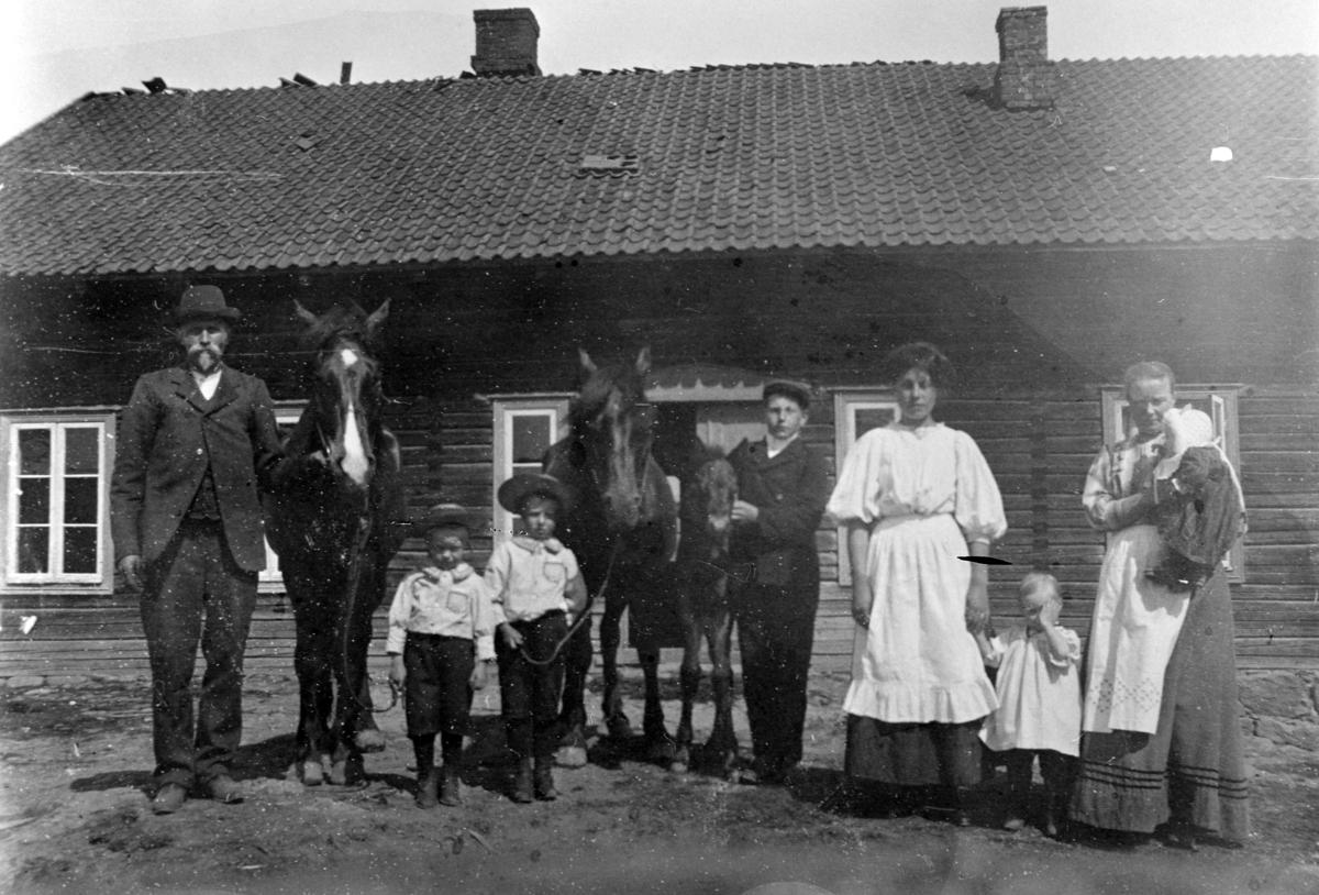 Gruppe mennesker oppstillt utenfor hus. Syverud, Ringsaker. Fra venstre er Lars Fossum f.1875, Leonard Fossum f.1906, Hans Østbye f.1905, Johan Melby f.1896, ukjent, Karen Fossum f.1909 , Gunda Fossum f.1874 med Dortha på armen. Hest med føll.