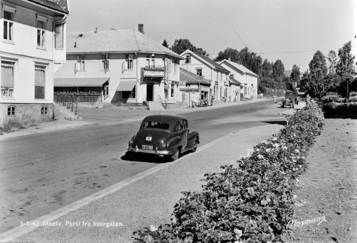 Postkort, Ringsaker, Moelv sentrum, Storgata 114 til venstre Apotekergården med M. P. Dobloug, Moelv Gjestgiveri, gullsmed Alf R. Haugen, slakter Arne Nygaard kjøttbutikk, Bjørges isenkram, Moelv bokhandel, Moelv fargehandel, bil med registreringsnummer A-60937,