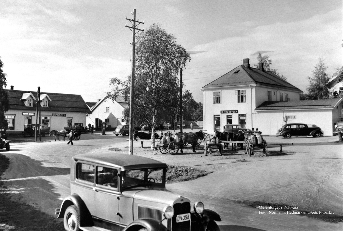 Postkort, Stange sentrum, Stangebyen, fra venstre: Magnus Godagers forretning i Storgata, og O, Nordsveens butikk til høyre, hestebinding på meieritorget, hester med stuttkjerrer kjører mjølk, en bil: Ford i forgrunnen med registreringsnummer D-4358, i heftet Hedmarks Bileiere 1938 er bilen registrert på Ole Wie,