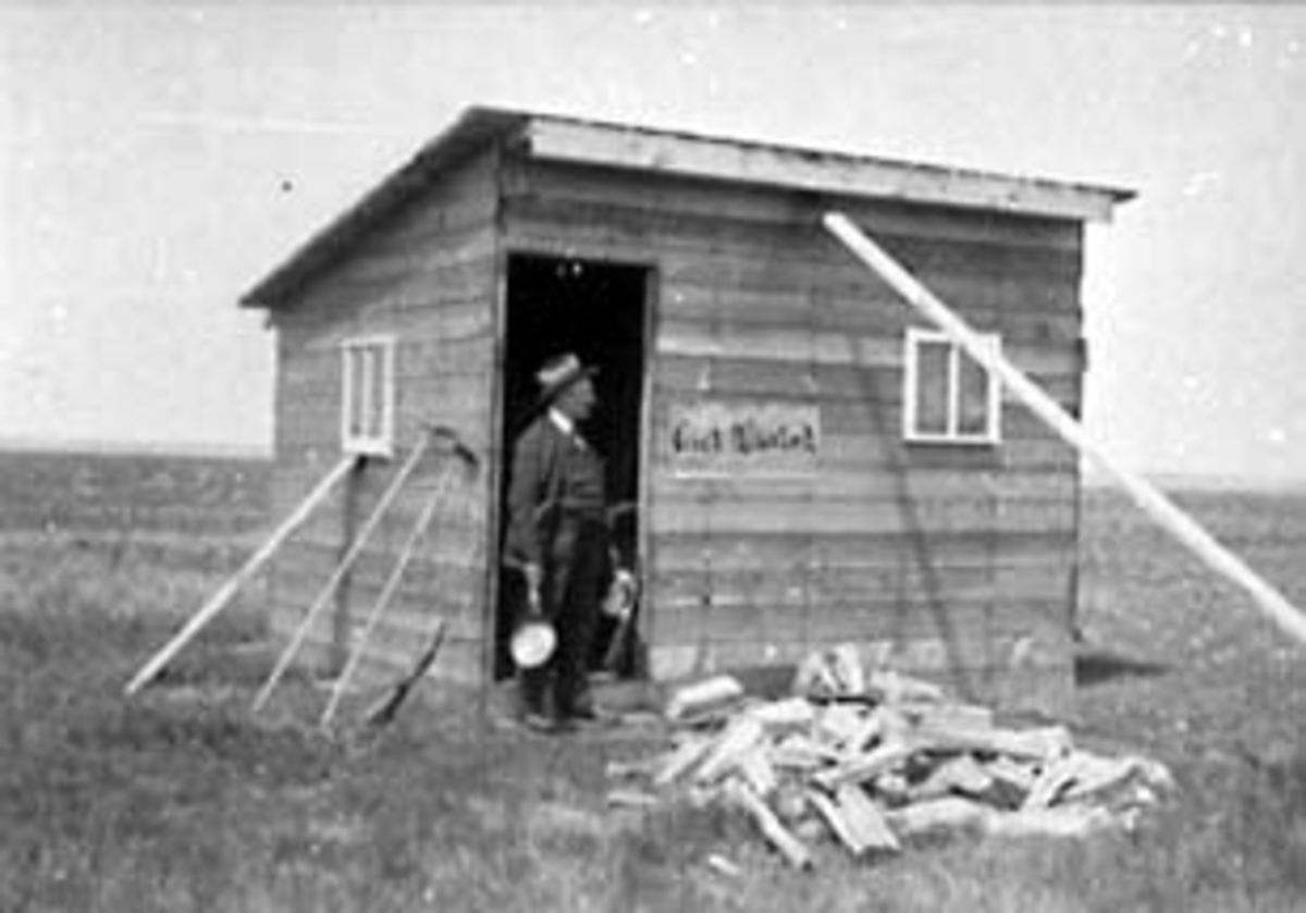 EKSTERIØR: BOLIG, "SHANTY", PLAKAT PÅ VEGGEN: "GIRL WANTED", MANN I DØRA, USA, UTVANDRER