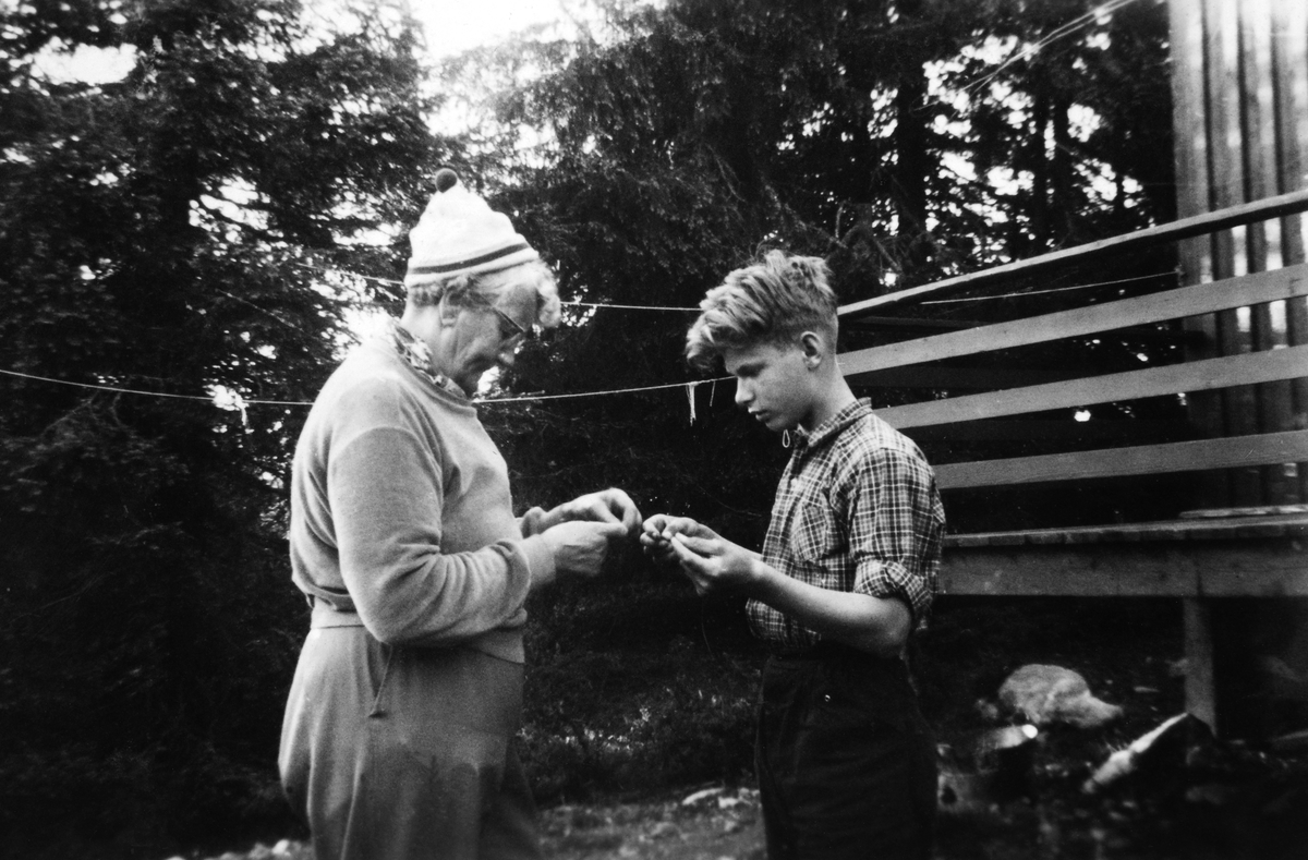 Harda M. Oppegaard viser Odd Skjøthaug hvordan en skal feste fiskekrok og flue til sene utenfor salmaker Arild Lundbergs hytte på Vassbakken ved Sjusjøen, juli 1955. 
Se artikkel i "Gammalt frå Stange og Romedal 2010" utgitt av Stange historielag. 