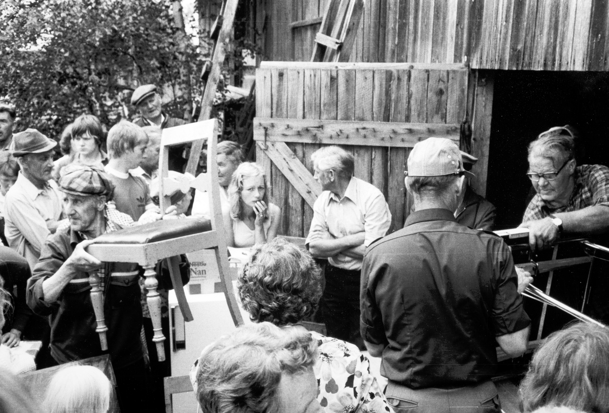 Auksjon ved Sæterskverna 12. juni 1980. peder Rønningsbakken lemper en biedermeierstol videre til kjøper. Med ryggen til i mørk skjorte auksjonarius lensmann Stein Huse og i døråpningen skraphandler Magnus Myra sen. 