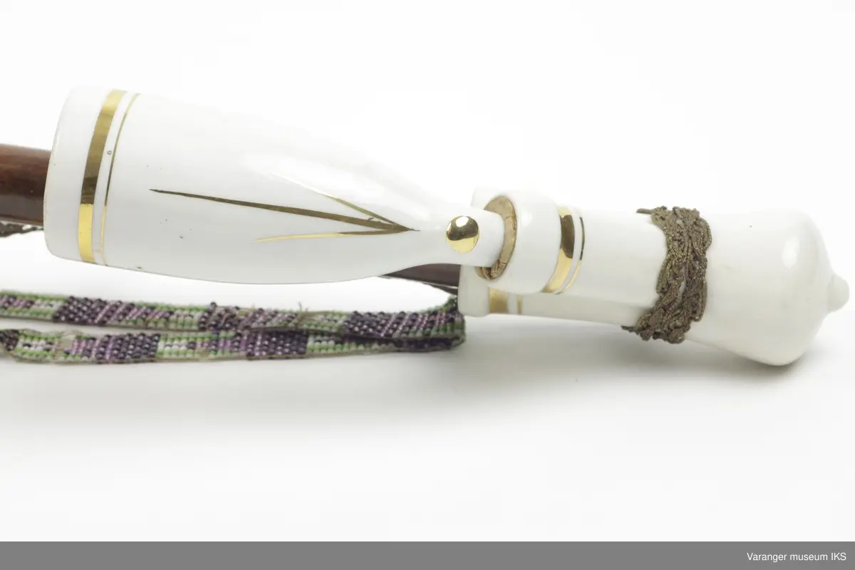 Lang, krum pipe. Hode av hvitglasert porselen med gullstriper. Skaft av mørk brunt tre. Den krumme delen av lær delt med flettede svarte stive hår og med enestykker av ibenholt. Munnstykke av bein, pyntet med vulster. To lange snorer, går fra øverst til nederst på det lange trestykket.