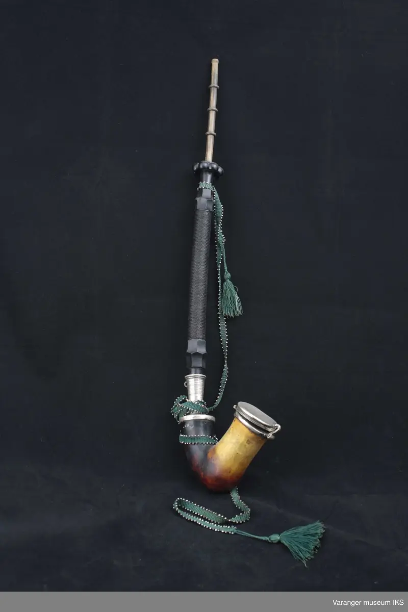 Lang, rettskaftet pipe. Hode av merskum med lokk og overgang til skaft av sølv. Skaft av lær med svart fletting rundt og med endestykker av ibenholt. Munnstykke av ben med vulster. På sølvlokket inngravert "H.E.Erindring fra 1882." Grønt vevet bånd med dusker og innvevete metallperler. Skaftet ser ut  til å høre til en annen pipe opprinnelig, da det er gjenger på det stykket som settes fast i sølvtuten.