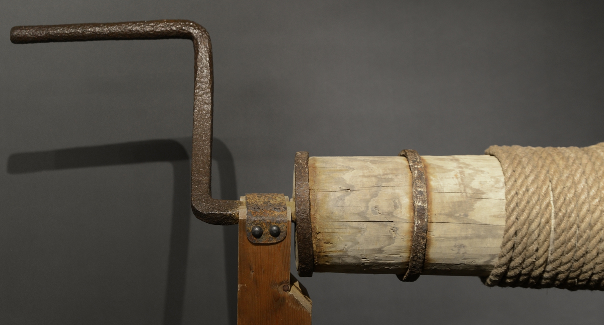 Heiseanretning av tre med sveiver i jern. Tauverk er festet til en vinsj som er montert i taket i utstillingen.