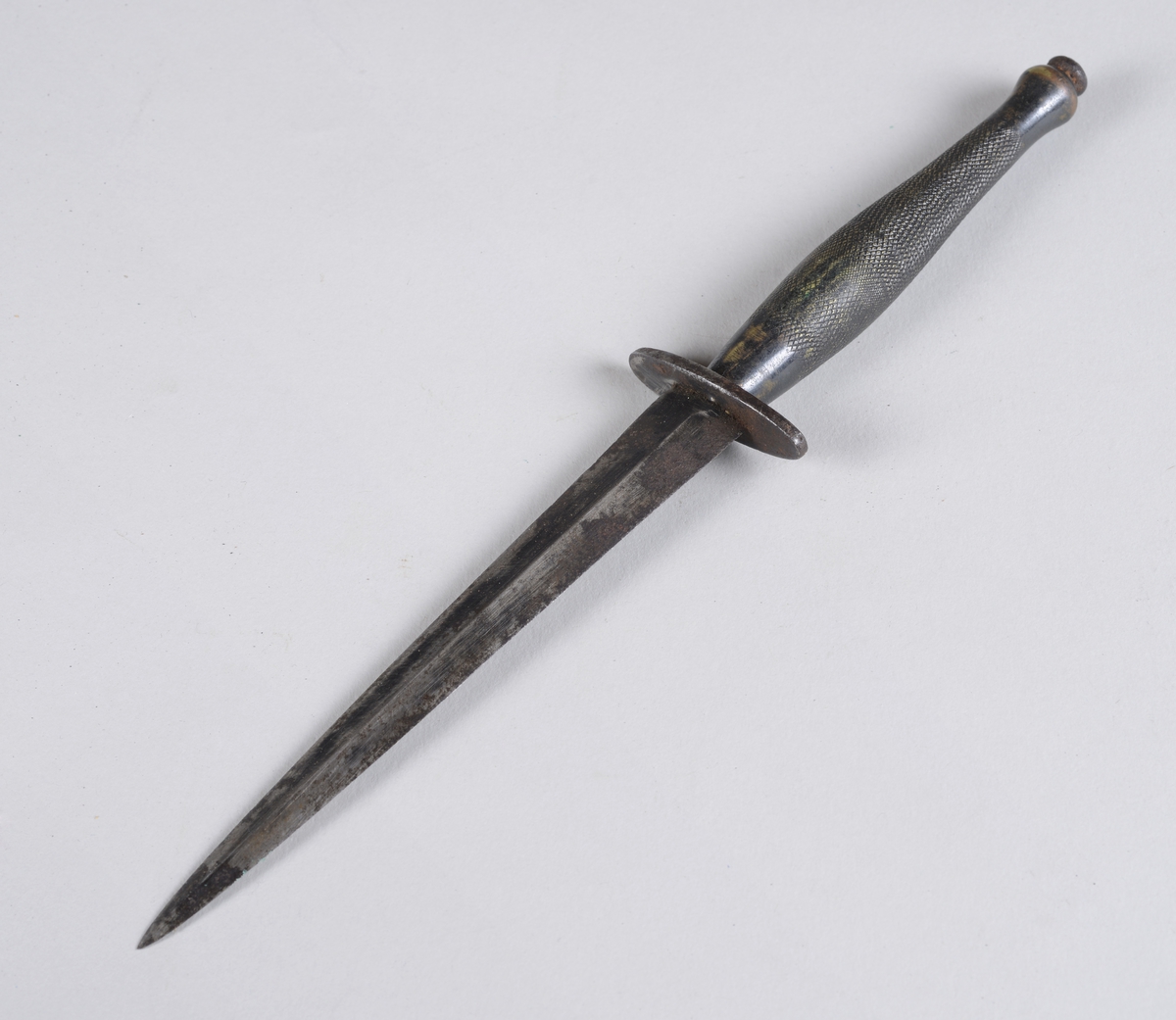 Dolk av jern i lærhylster. 
Hylsteret er forsterket i spissen og kan festes i belte. Dolken har et ruglete håndtak og et toegget blad med midtribbe.