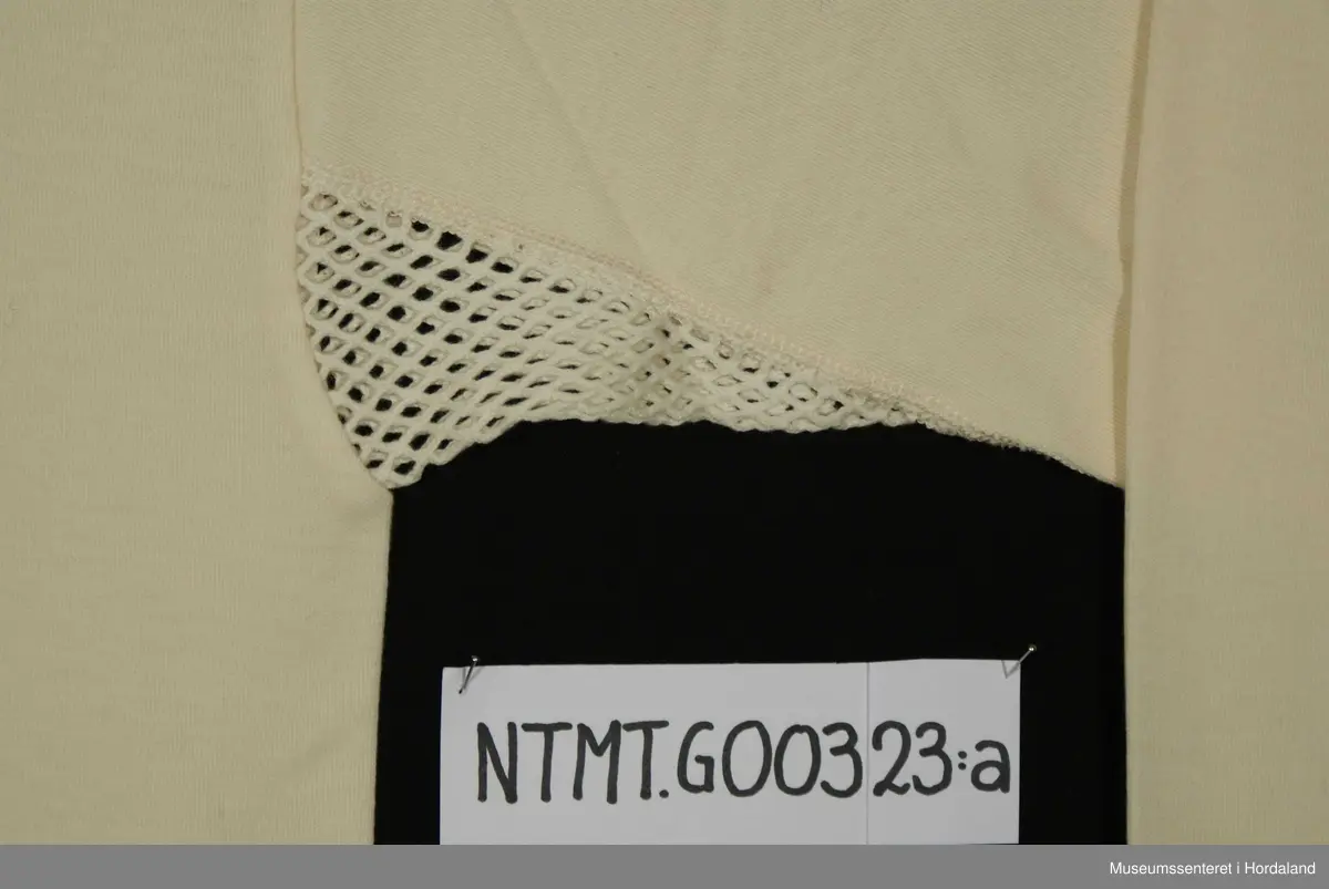Rund hals med åpning midt foran med to knapper, lange ermer med brotavslutning, kile under ermer i helsetrøyestoff, skulderstykker.