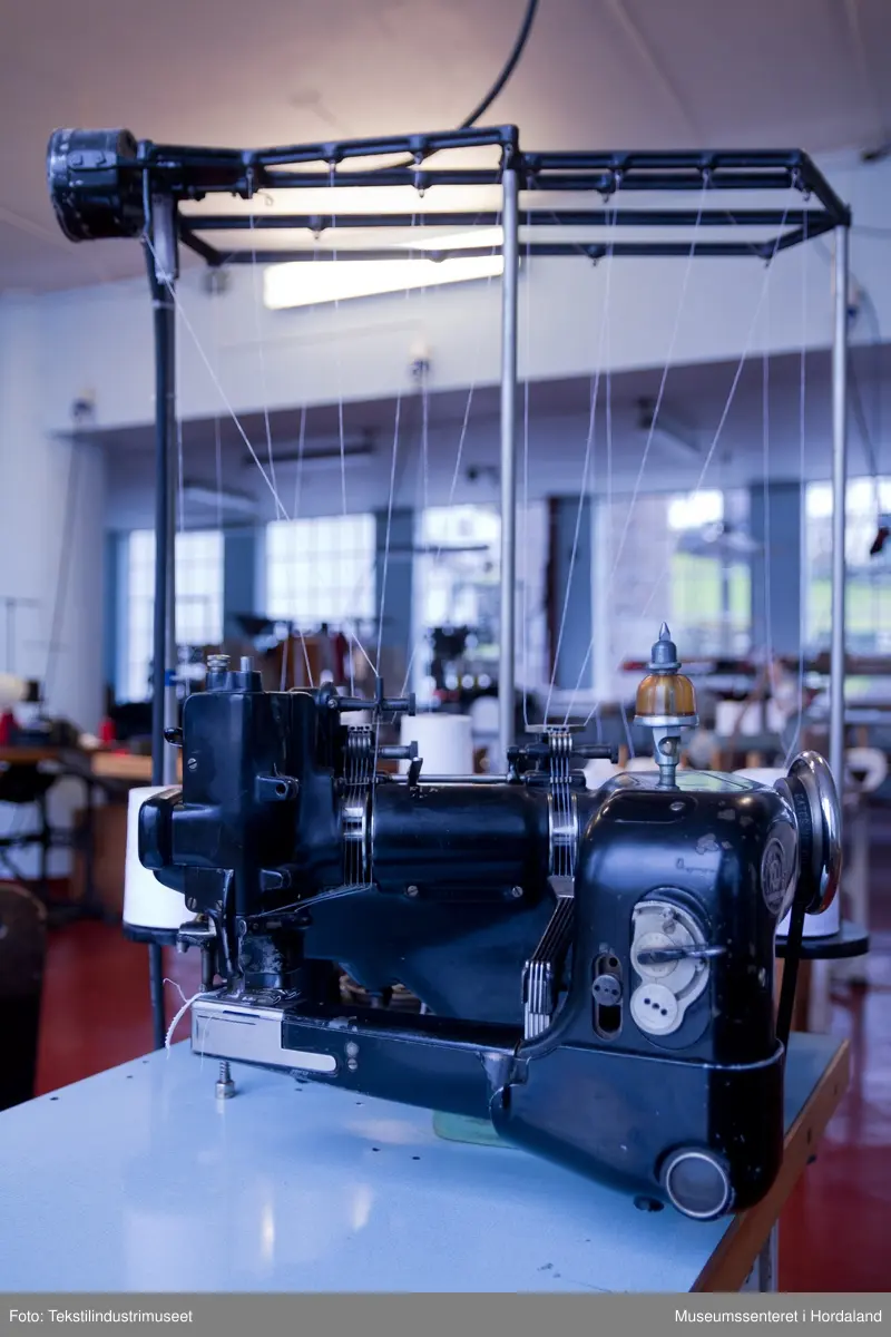 Flatlock-symaskin i svartmåla støypejern og med halvautomatisk oljing. Maskina er montert på eit Juc-bord og har Singer-motor (truleg ikkje original).

Motor: Singer alternating current electric transmitter. No. K 692357 R
Volts: 210-230.