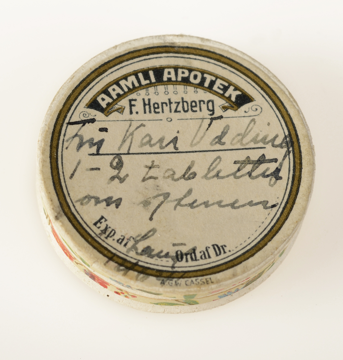 Rund pilleske laget av papp med dekorativt papir på sidene. Lokket og bunnen er omtrent like store, lokket kan tas helt av. På lokket er apotekets signatur (etikett).