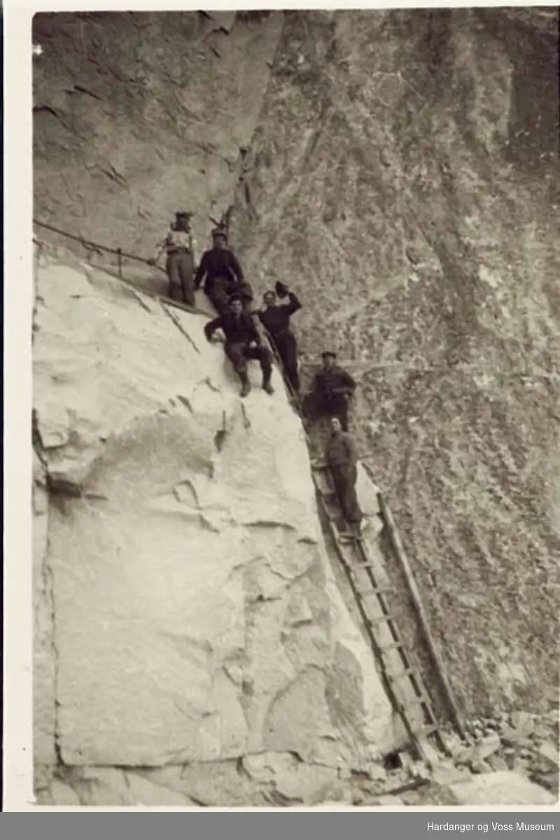 Gruppe, menn, fjellnakke, stige, vegarbeid. Dei sit på nakken og står i stige. Ved Langfoss