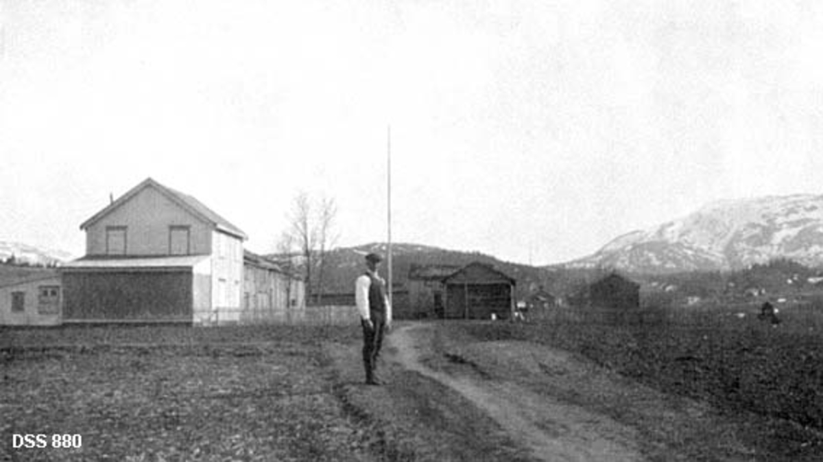 Garden Nordre Forsjord i Vefsn.  Fotografiet er tatt fra gardsvegen, der det står en mann med kvit skjorte, vest og sixpencelue.  Bakenfor ham ses bygningene.  Til venstre ligger det ei rekke våningshus.  Det nærmeste virker forholdsvis nytt, er bordkledd og malt i en lys farge, med et tilbygd enetasjes skur med pulttak ved gavlen.  Bakenfor skimtes et eldre, langt våningshus.  En bygning til høyre i tunet kaltes «Skoghuset».  Et annet gardstun skimtes til høyre, bakenfor Nordre Forsjord.  I bakgrunnen åsrygger og fjell som delvis er snødekte.