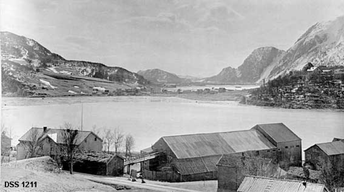 Landskapsfotografi fra Kulstadsjøen i Vefsn på Helgeland mot Halsøy og Mosjøen.  Fotografiet er tatt fra en bakkekam med et gardsbruk i forgrunnen.  Her skal det også ha vært en liten bedrift, Nils Skandfers båtbyggeri.  På motsatt side av den nærmeste sjøflata er det til venstre lier med skog- og engarealer, til høyre et berg med enkelte store bygninger.  På eidet midt på bildet er det reist en del hus.  Bakenfor skimtes ei sjøflate med pælerekker, antakelig til tømmerreservoaret for Nesbruket.  Bakenfor dette igjen skimtes tettstedet Mosjøen og ei markant, bratt fjellrekke (Øyfjellet). 