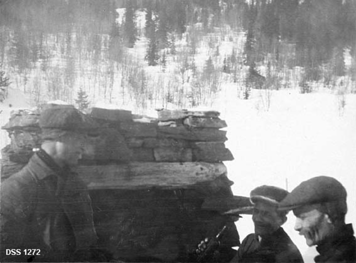 Tre menn på Nes trelastbruks eiendom Sirijord. Fotografiet er tatt vinterstid. Mennene, iført skyggeluer med ørelapper, står foran en murt natursteinskonstruksjon, muligens et ildsted som mangler omgivende bygningskonstruksjon. Den ene av herrene holder ei mørk glassflaske med drikke i handa. I bakgrunnen en snødekt landskap. Mangelen på vegeasjon som stikker opp av snøen gir grunnlag for å anta at det er dyrket mark akkurat der karene står, men i lia bakenfor vokser det bjørkekratt og en de spredte graner.