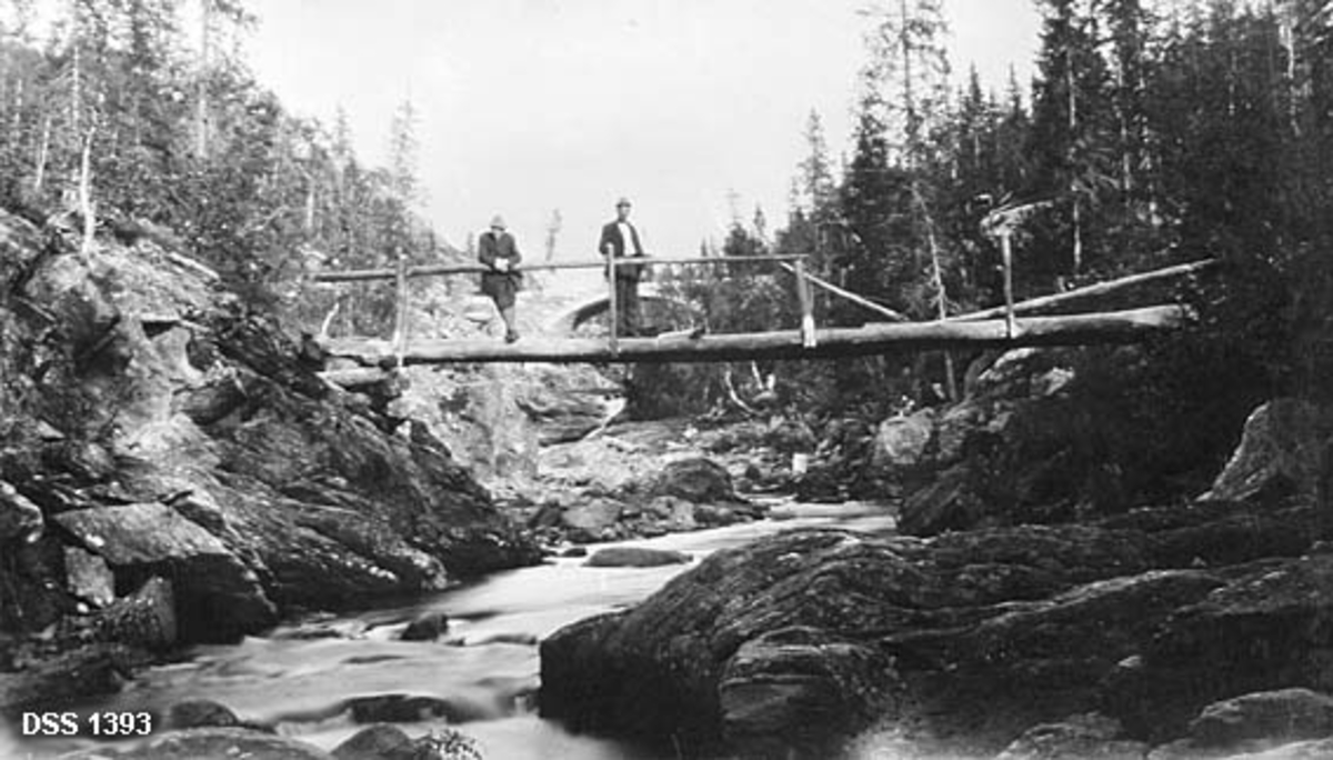 Bruer over Brekkvasselva i Brekken statsskog i Nord-Trøndelag.  Fotografiet er tatt fra elveløpet.  I forgrunnen ses ei einkel trebru, båret av tømmerstokker som er lagt slik at de danner et spenn tvers over elveleiet mellom to bergnabber.  Brua har enkelt trerekkverk som er delvis intakt.  To menn står på denne brua.  I bakgrunnen skimtes ei ny hvelvet steinbru, bygd for den nye hovedvegen fra Grong mot Nordland (jfr. DSS. 1392).  Landskapet langs elva mellom bruene har en del granskog. 