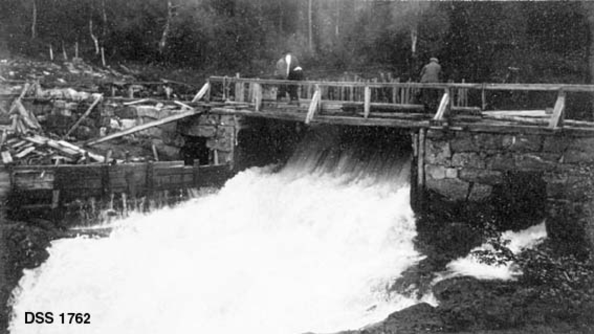 Lillebudammen i Trevja, sett fra motstrøms side.  Damarmene er oppført av tørrmurt gråstein.  Over damåpningen er det bygd ei trebru, der tre karere arbeider med å sette stengsel av nåler.  Til venstre for damløpet er det en plankeskjerm. 